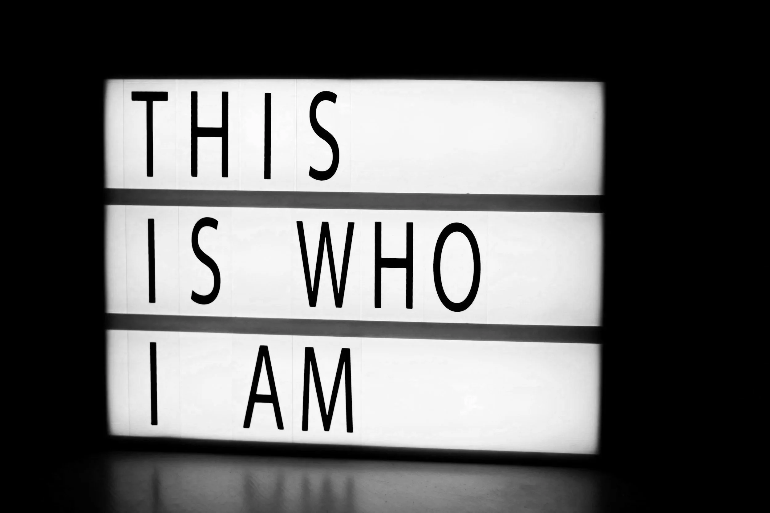 Lightbox sign with the words 'THIS IS WHO I AM' in black letters, with the text divided into three lines, on a white background, surrounded by darkness.
