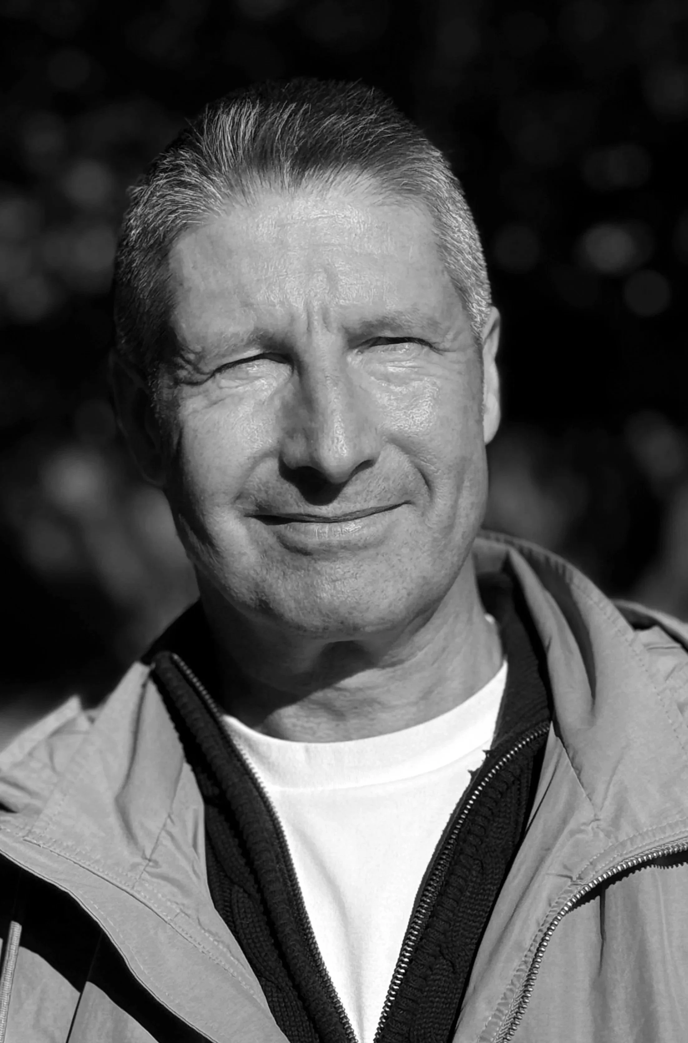 A black-and-white photograph of a middle-aged man with short, combed-back hair, smiling outdoors with a blurred natural background.