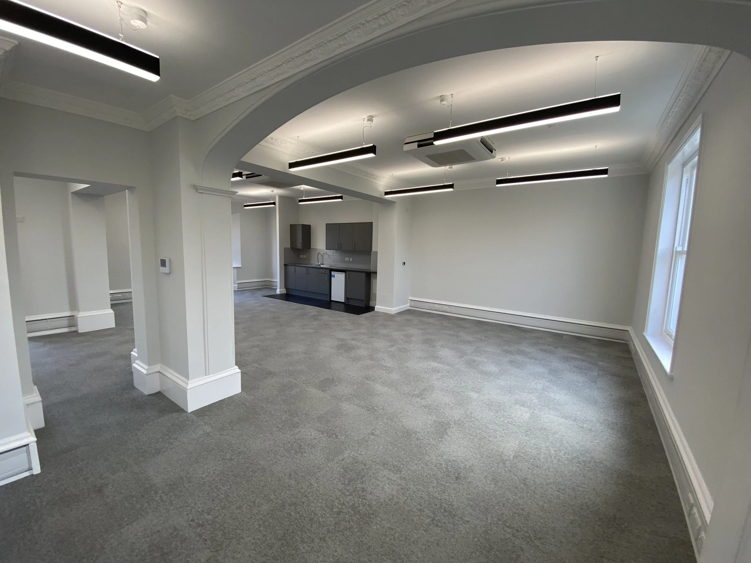 Empty room with gray carpet, white walls, and large windows, with a kitchenette and modern black lighting fixtures on the ceiling.