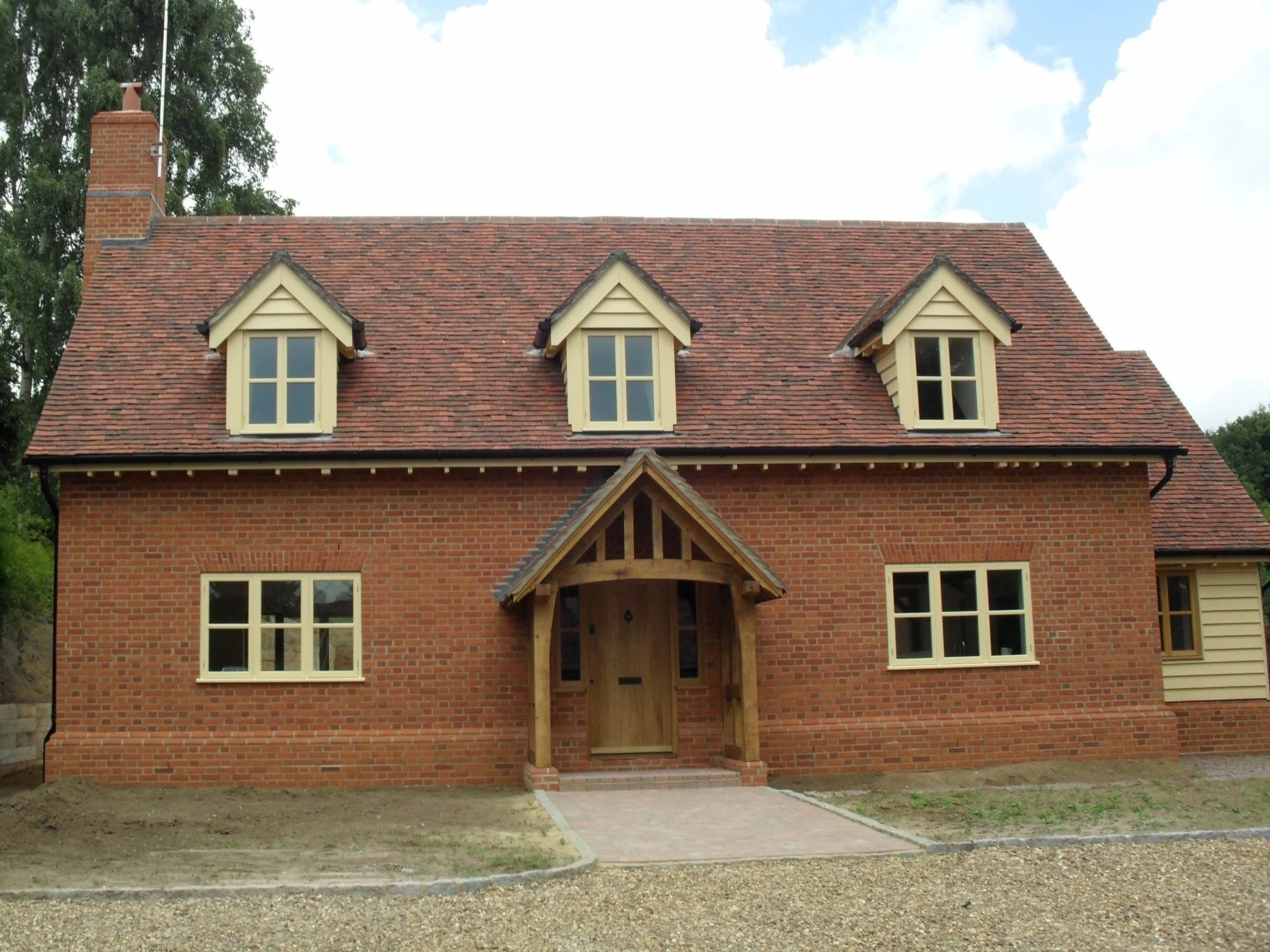 A brick two-story house with a steep red tile roof, three dormer windows on the upper floor, and four windows on the lower floor. The house has a small front porch with a wooden door and a gabled roof supported by wooden posts.