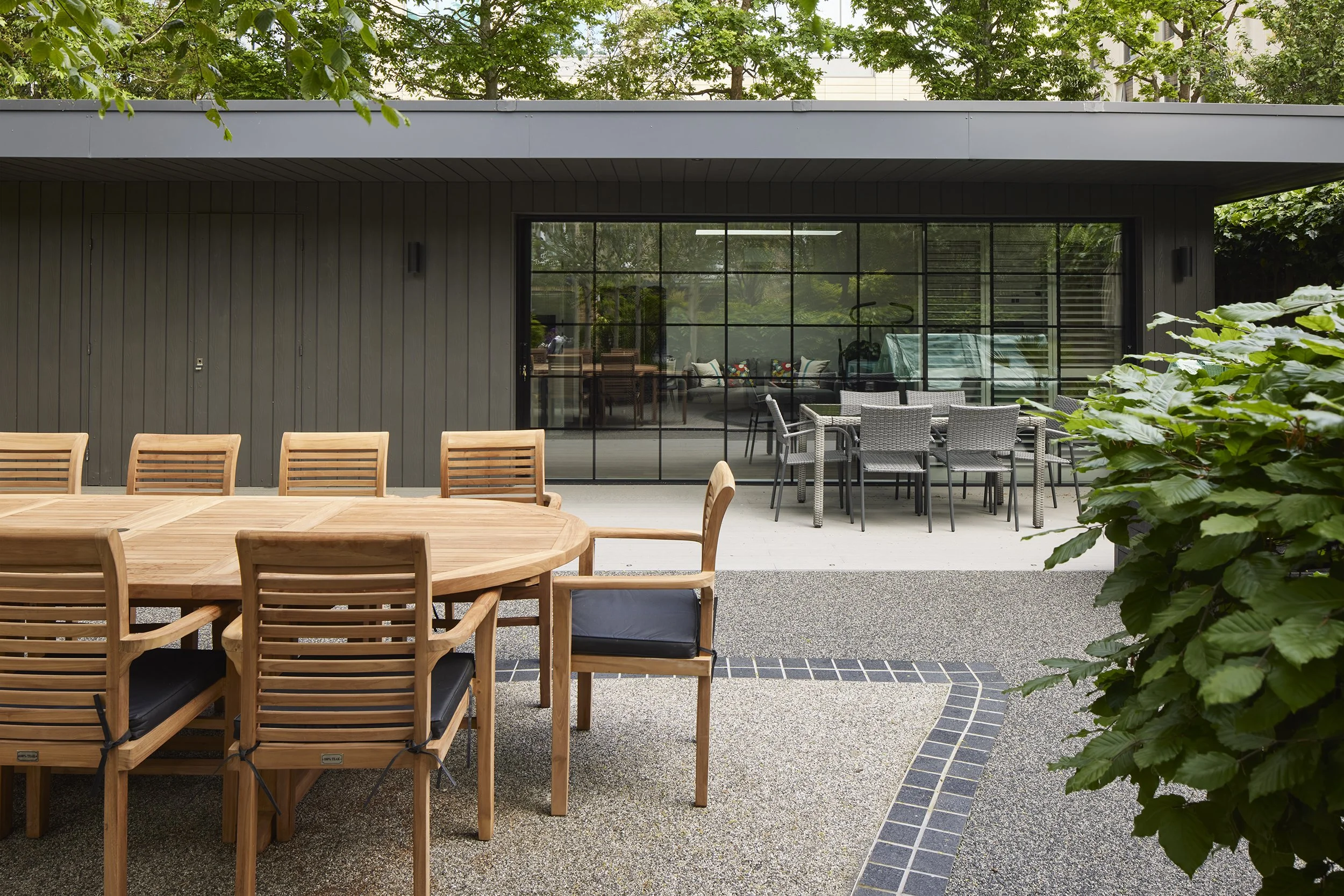 Outdoor patio scene with wooden dining table and chairs, and a separate set of gray wicker tables and chairs. A glass sliding door reveals an indoor living space with a blue swing and colorful pillows, surrounded by green trees and bushes.
