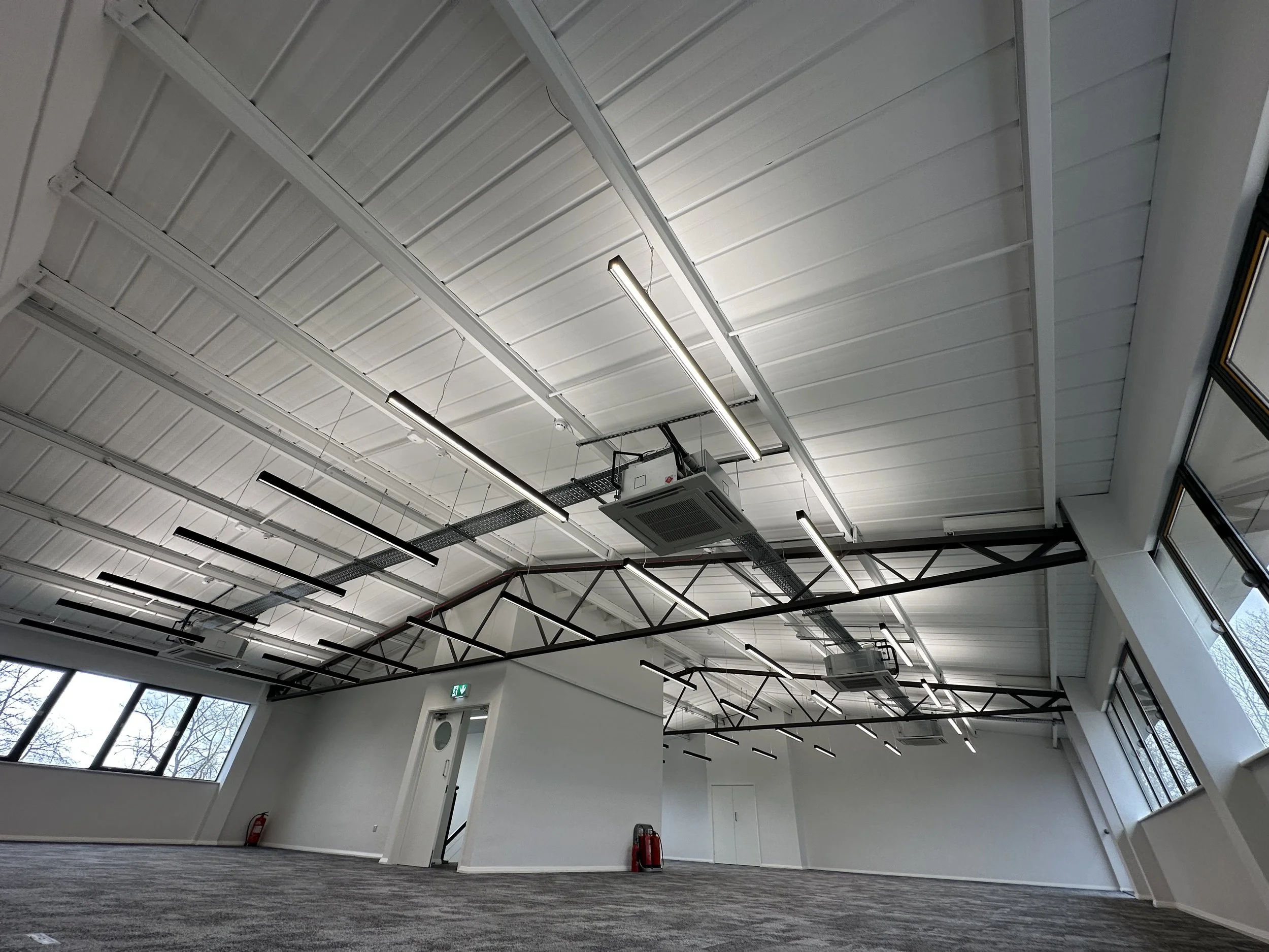 Empty commercial or industrial space with white walls, large windows, and ceiling with exposed beams, lighting, and ventilation units.