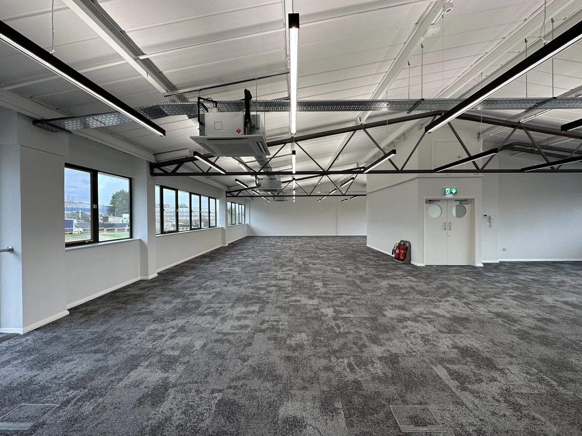 Empty office space with large windows, gray carpet, and ceiling-mounted lights and air conditioning units.