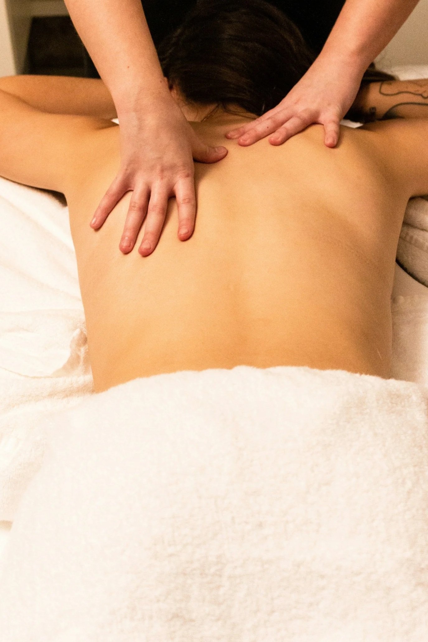 A person giving a massage to another person lying face down on a bed, with a towel partially covering their lower body.