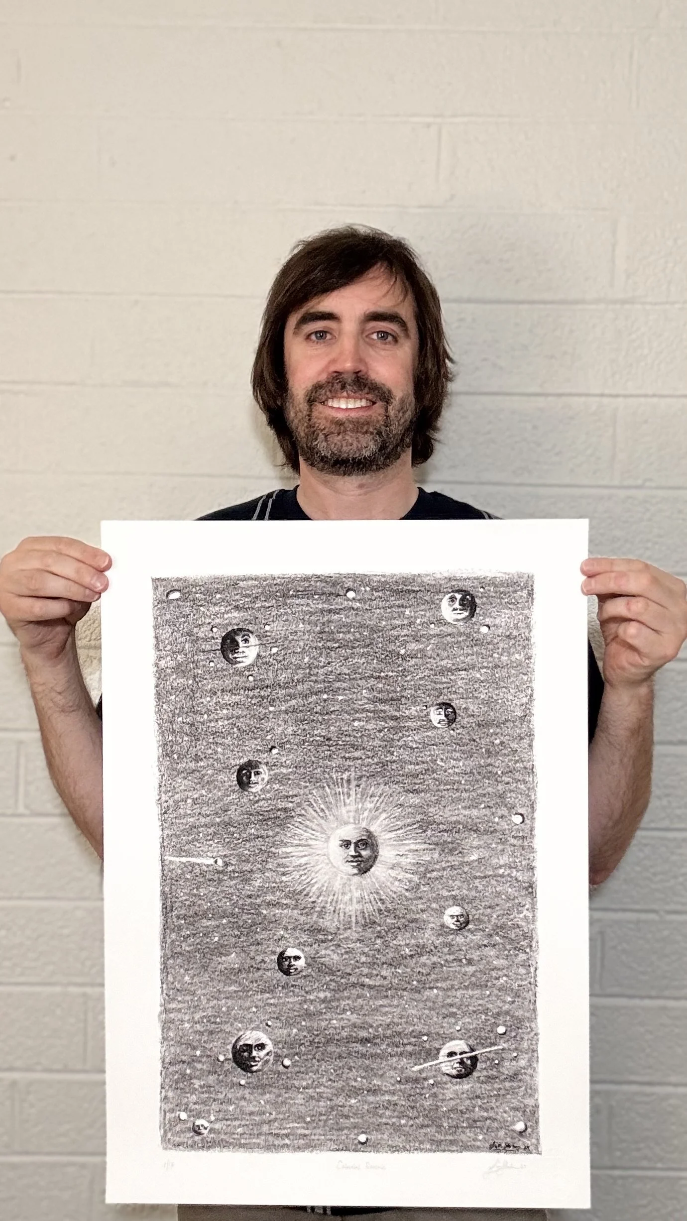 Artist Tim Stokes holding the limited edition print Celestial Reverie, a surreal charcoal drawing featuring celestial faces, moons, and cosmic symbolism.