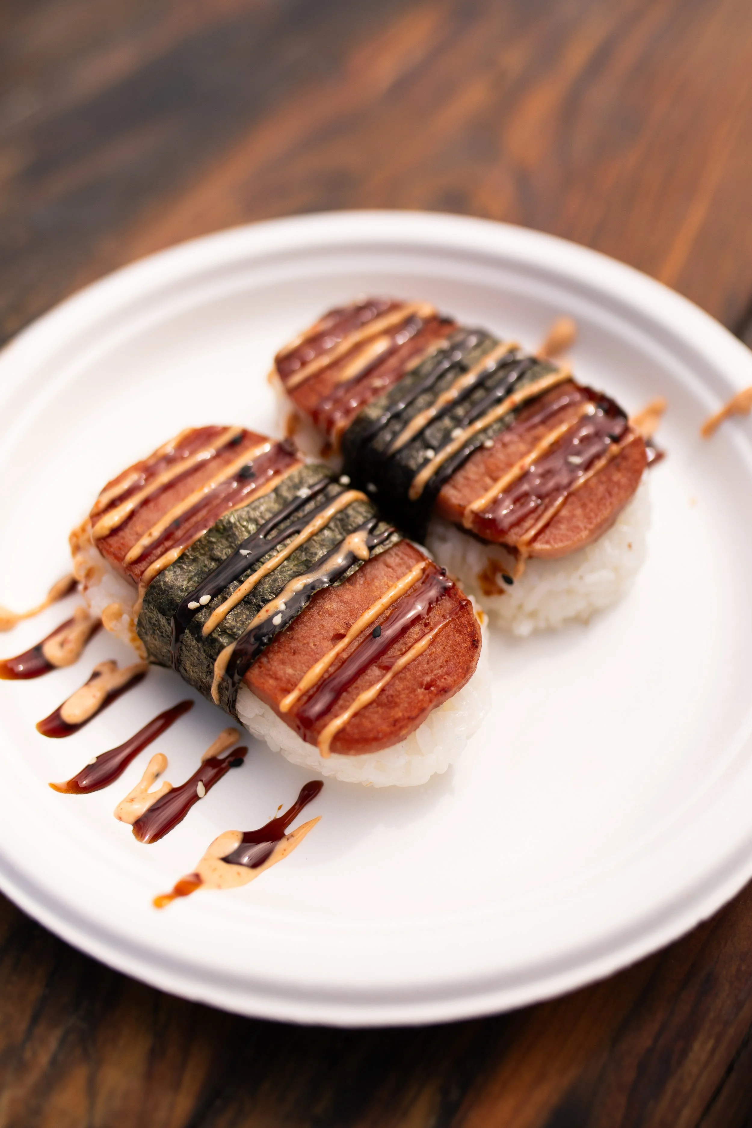 Two sushi rolls topped with sliced processed meat, drizzled with sauces, served on a white plate with a dark wood table background.