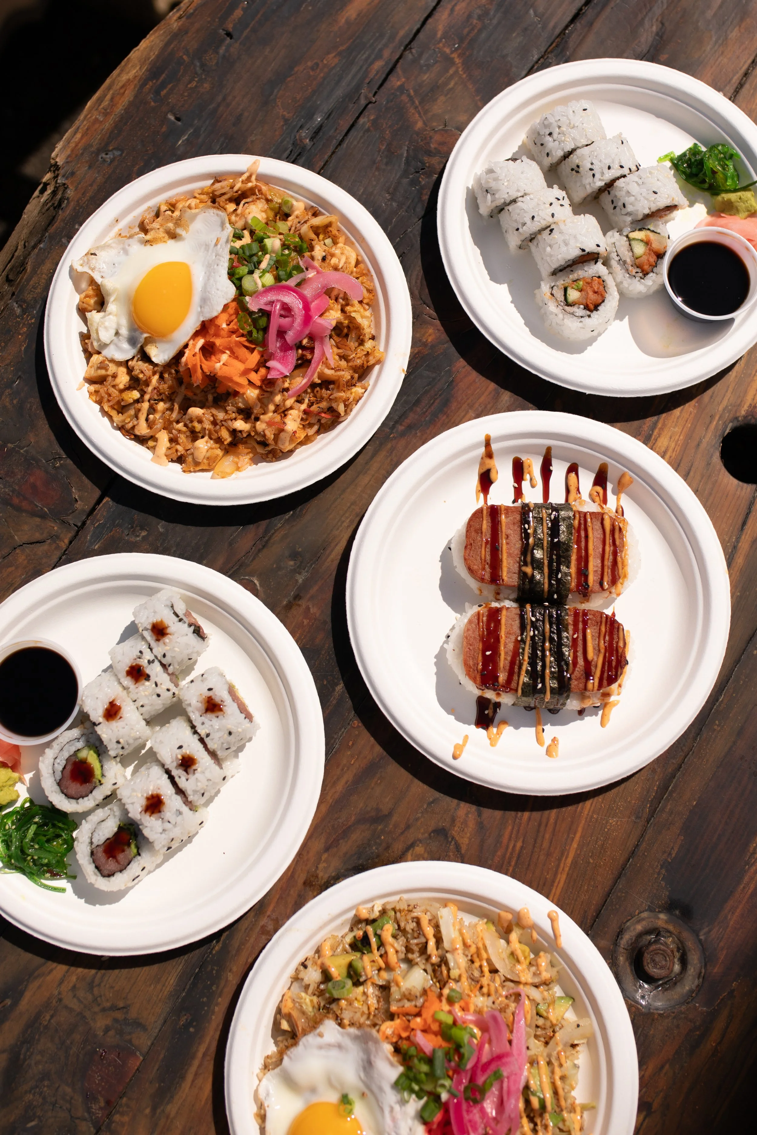 Assorted sushi rolls, rice dishes, and fried sushi with sauce served on white disposable plates on a wooden table.