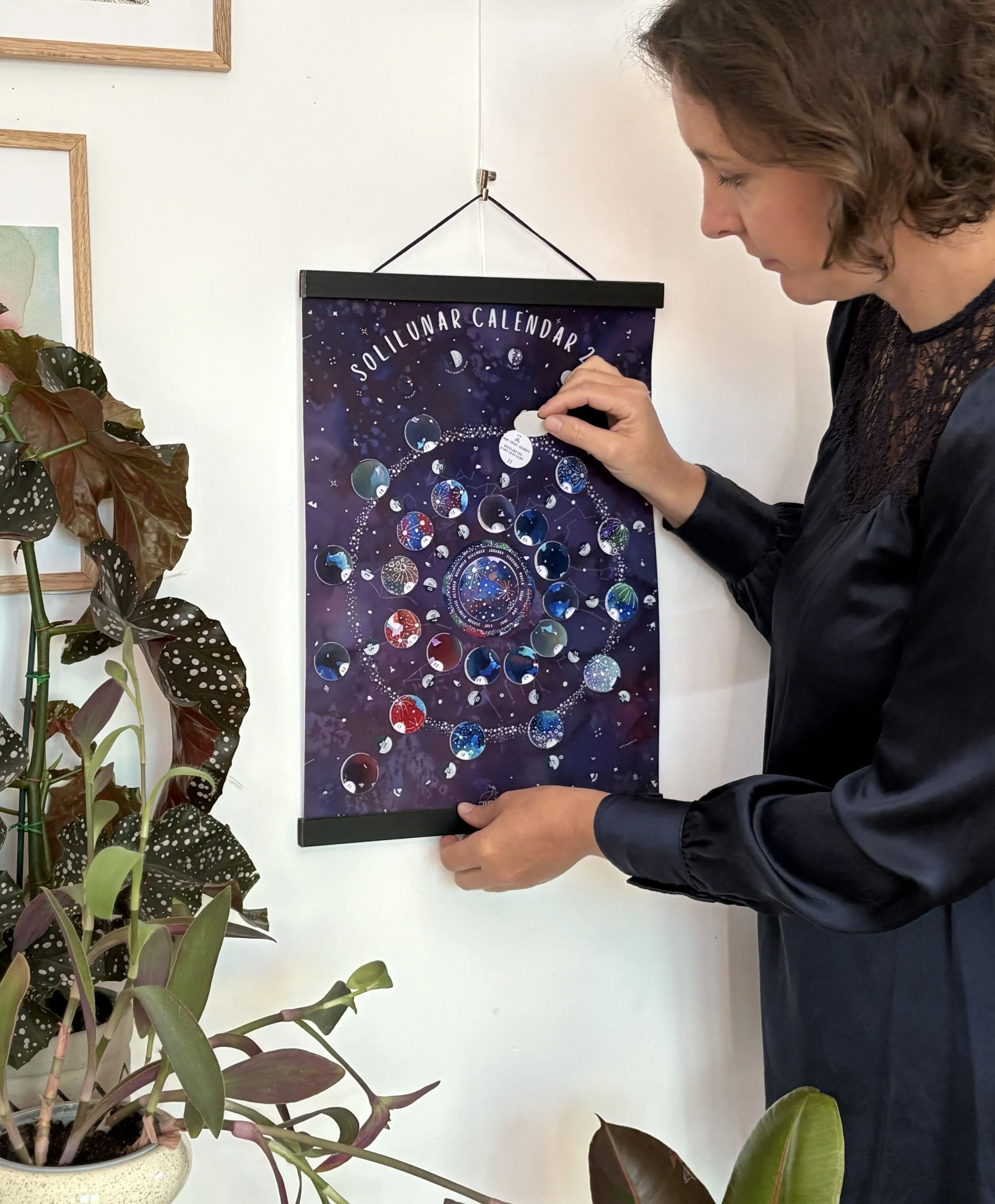 Full view of the solilunar calendar 2027, with a woman lifting an interactive flap on the 2027 Moon Calendar against a purple starry background.