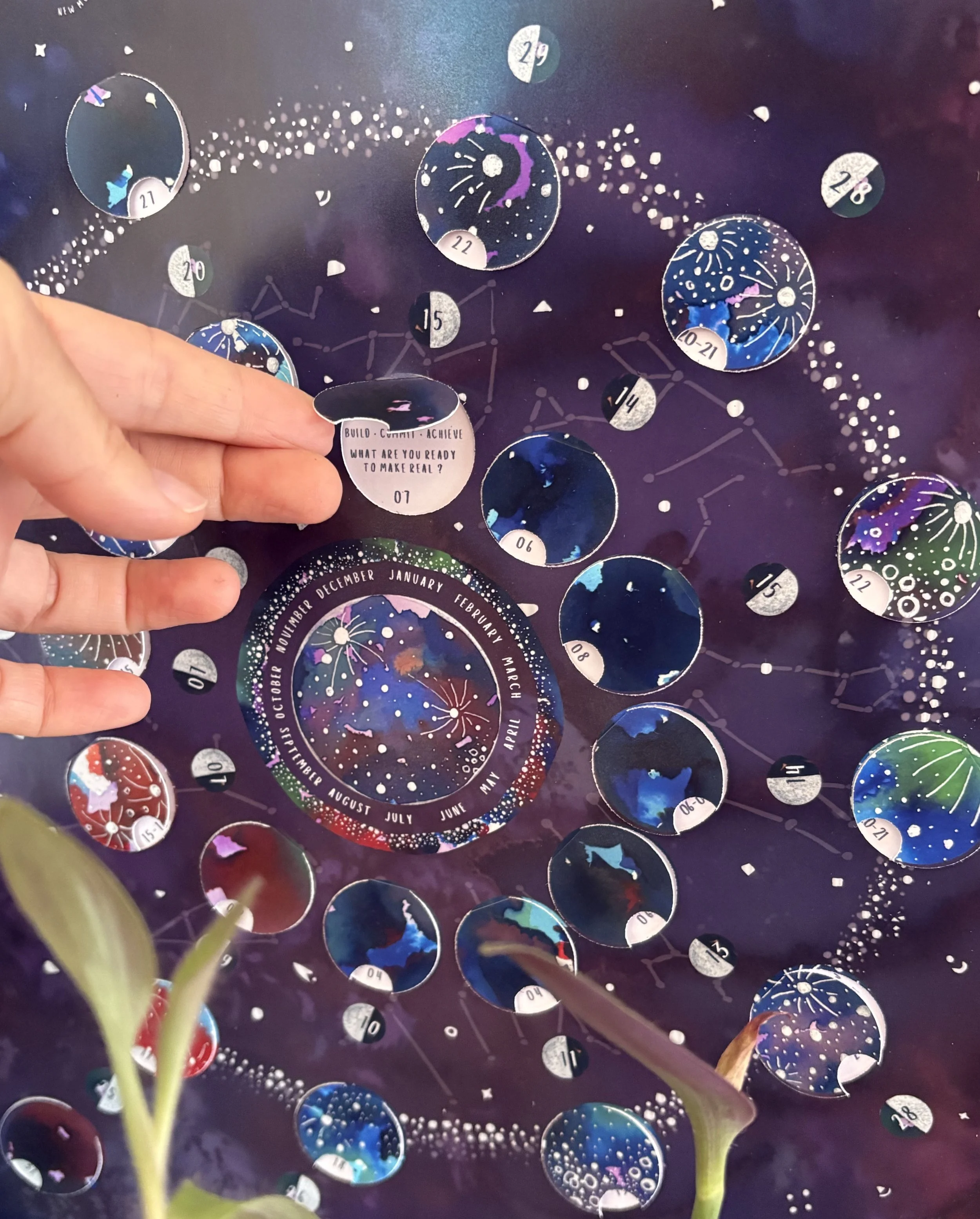 Close-up of a hand lifting an interactive flap on the 2027 Moon Calendar to reveal the Leo moon sign, three keywords and a reflection prompt, against a purple starry background.