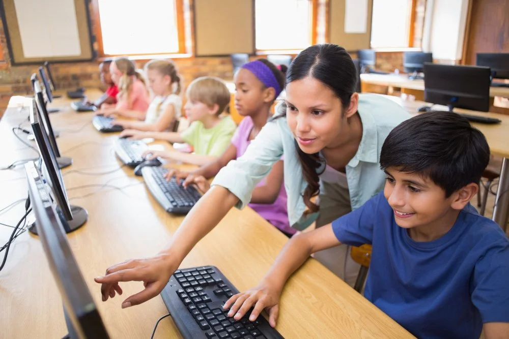 A female instructor guiding a young student on a computer in a classroom filled with diverse children learning tech skills.