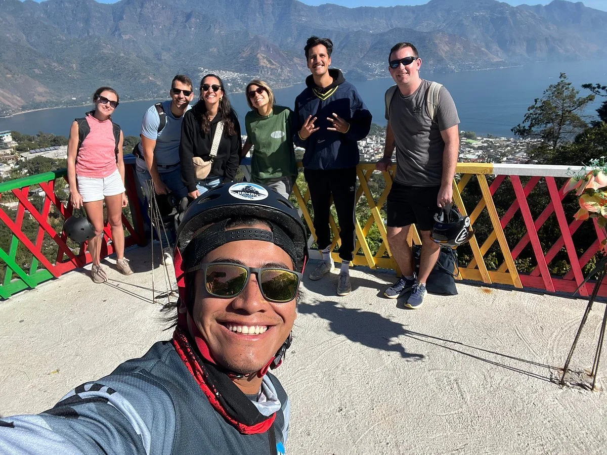 Smiling group of seven people on a scenic overlook with mountains and a lake in the background, some holding helmets.