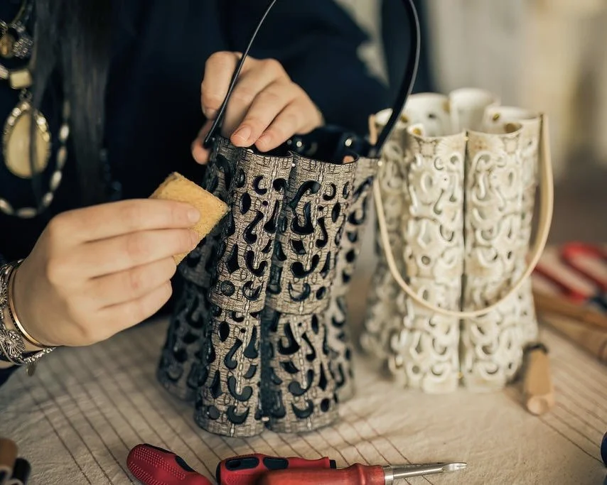 Close-up of designer Ana Del Rio's hands carefully finishing and cleaning a structured leather printed panelled handbag featuring intricate cut outs. This one is black and grey and a second version in cream and beige tones sits in the background.