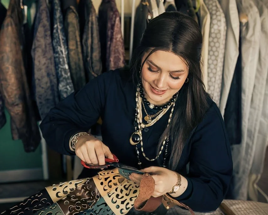 Close-up of designer Ana Del Rio carefully cutting a piece of leather of the strap of a decorative, panelled multi-coloured leather dress. Behind her, various garments hang on clothes rails in a studio.