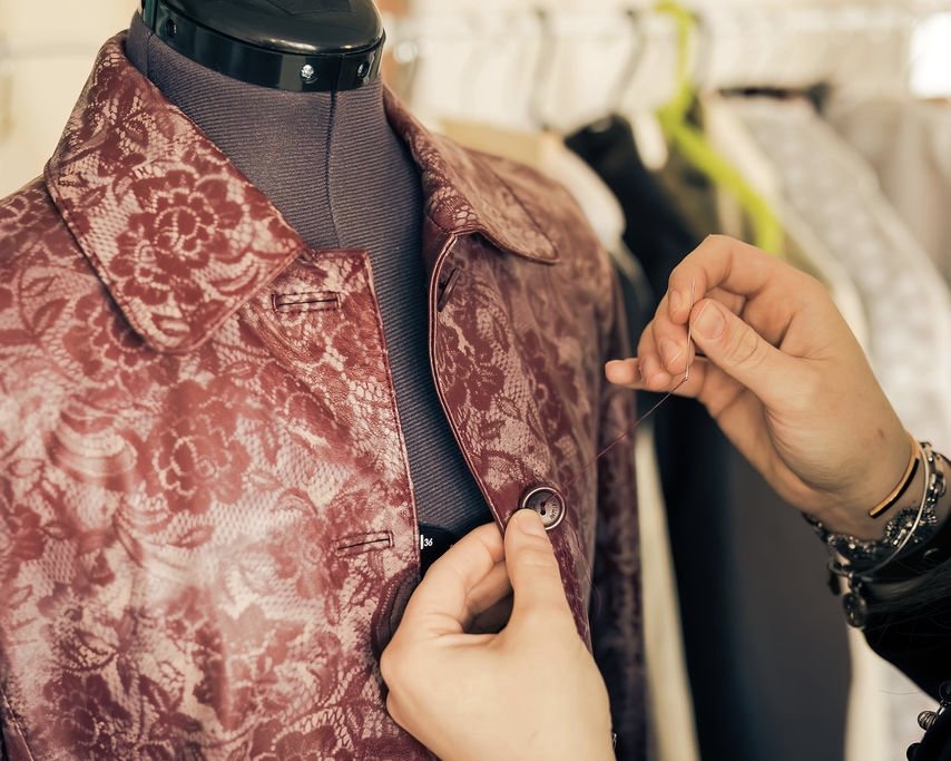 Close up of designer Ana Del Rio's hands hand-sewing a button onto a structured leather jacket with a red and cream floral print pattern, displayed on a grey mannequin.