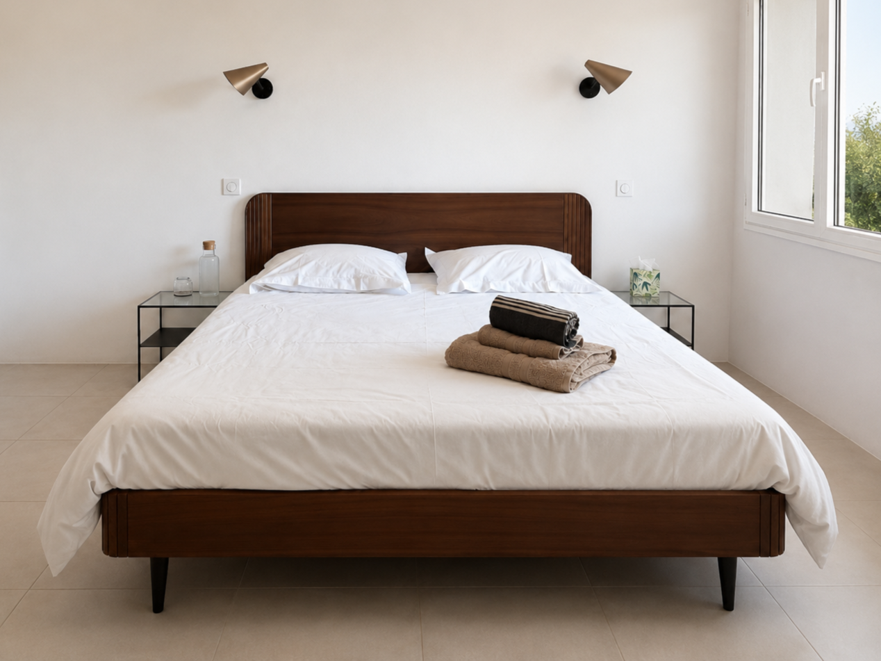 A neatly made bed with white linens and pillows, a wooden headboard, two black-and-white towels folded on top, with side tables holding a glass water bottle and tissue box, in a minimalistic bedroom with natural light from a window.