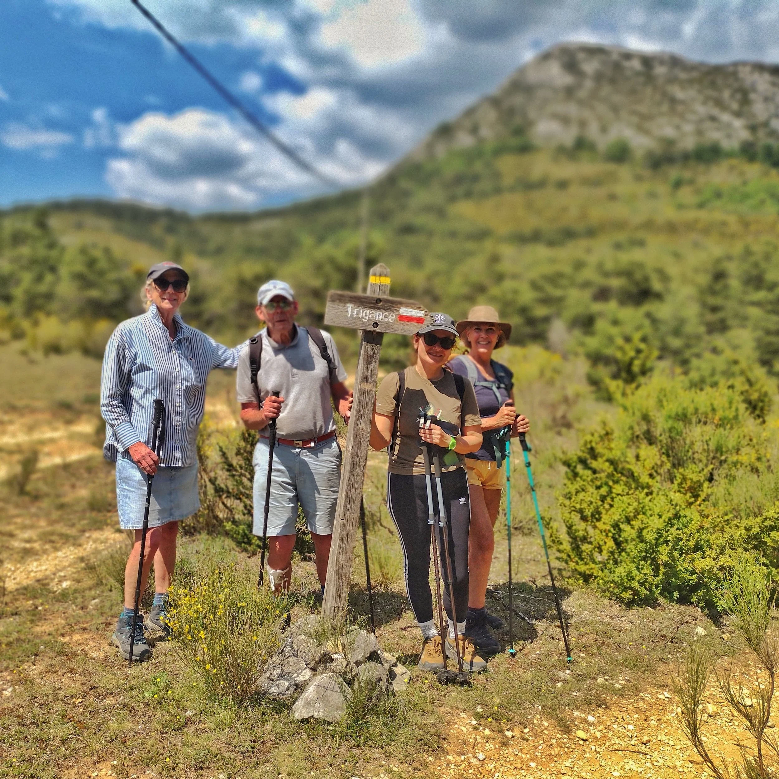 A group of hikers standing outdoors on a trail in a mountainous area, holding trekking poles, with a signpost indicating to Trigance, and a hill with a rocky summit in the background under a partly cloudy sky.