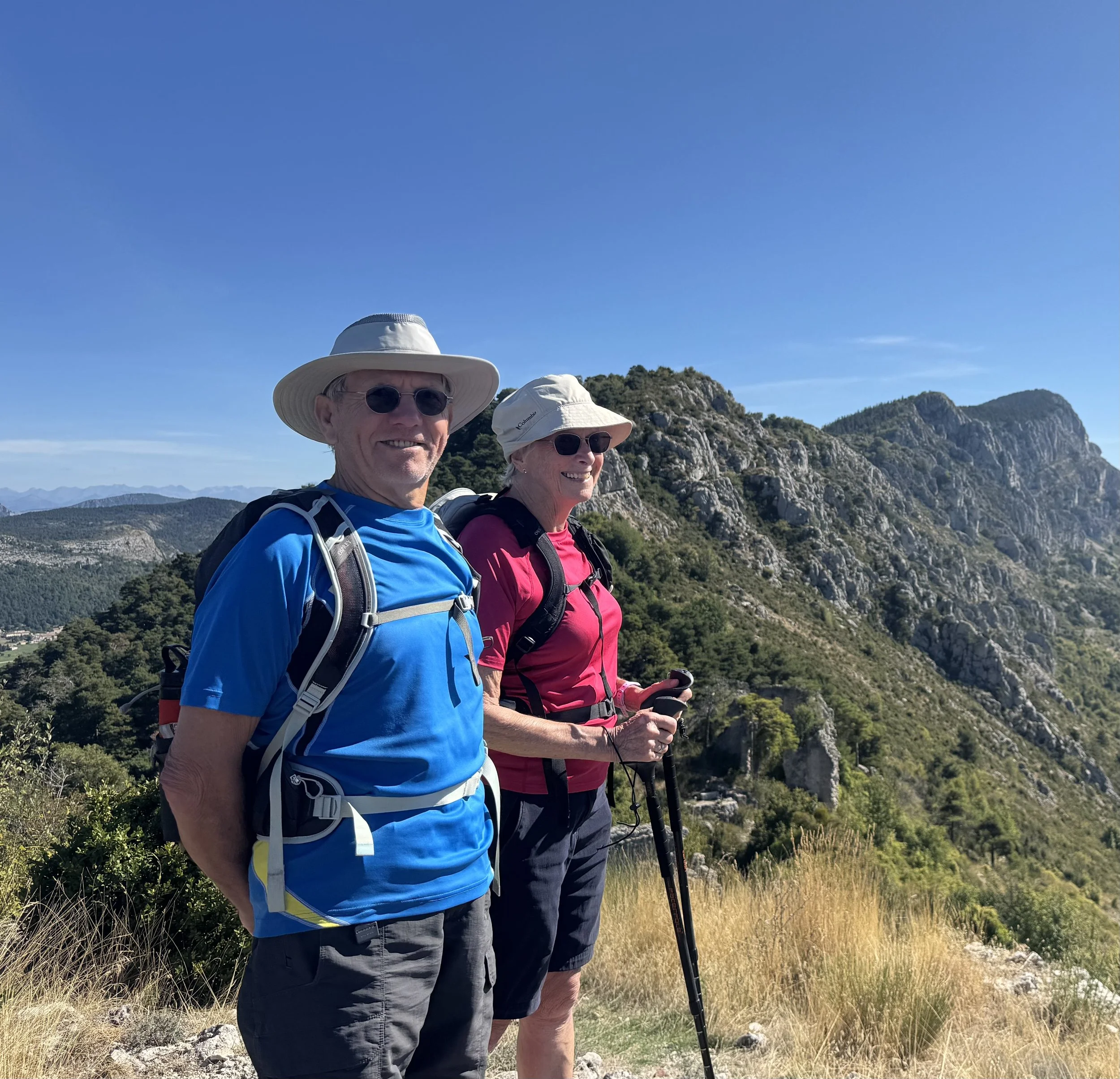 An senior man and woman hiking in a mountainous area, both wearing hats, sunglasses, and backpacks, with a rocky, green mountain landscape behind them under a clear blue sky.