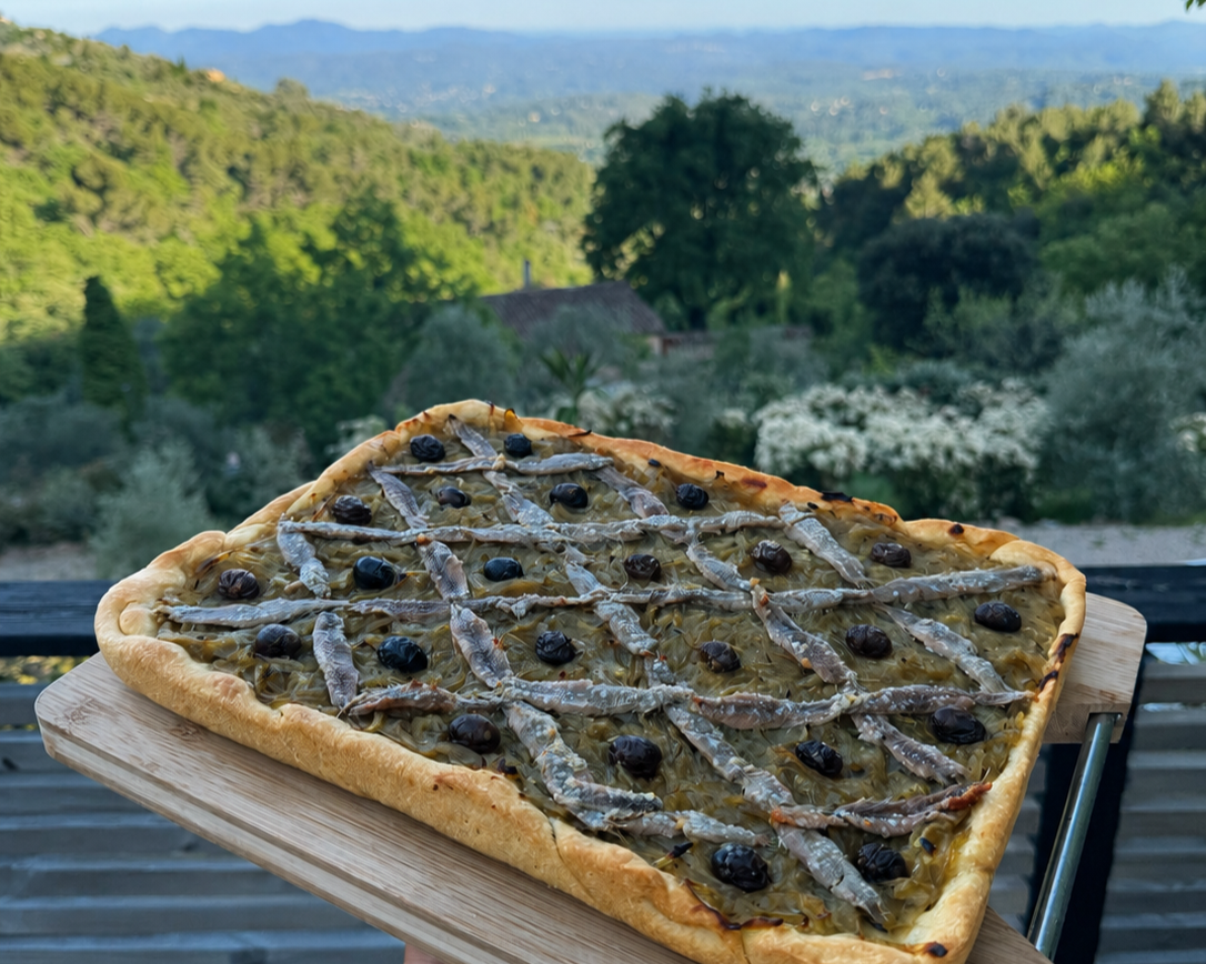 A rectangular pizza topped with small fish, black olives, and caramelized onions on a wooden board outdoor with a scenic view of trees and mountains in the background.
