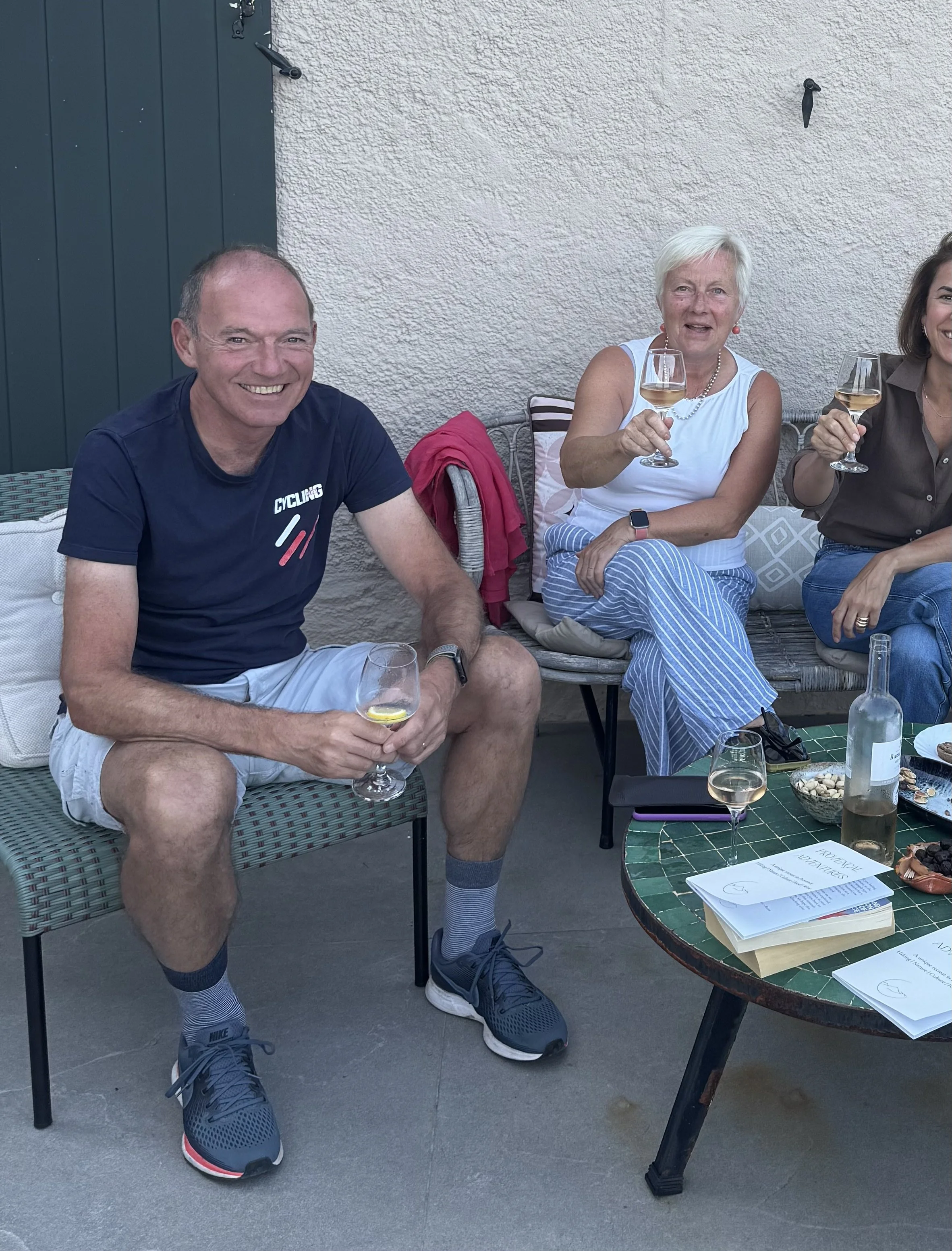 Three people sitting on outdoor benches, smiling, and holding glasses of white wine