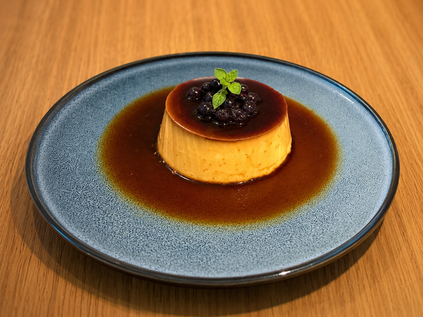 Caramel flan dessert garnished with blackberries and a mint leaf, served on a blue plate.