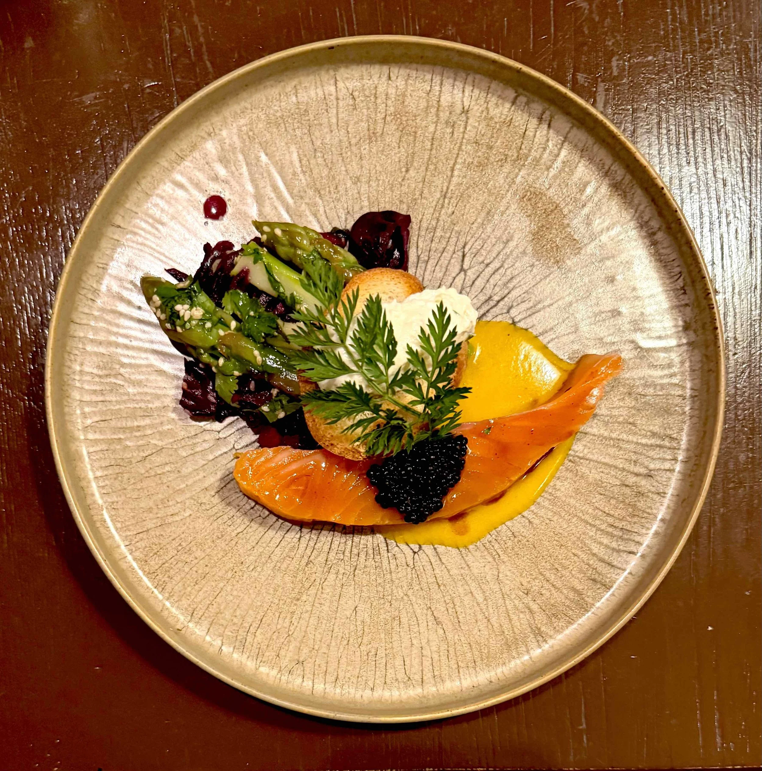A plated gourmet dish featuring smoked salmon, black caviar, mashed potatoes, fresh greens, and a sprig of herbs on a ceramic plate, placed on a wooden table.