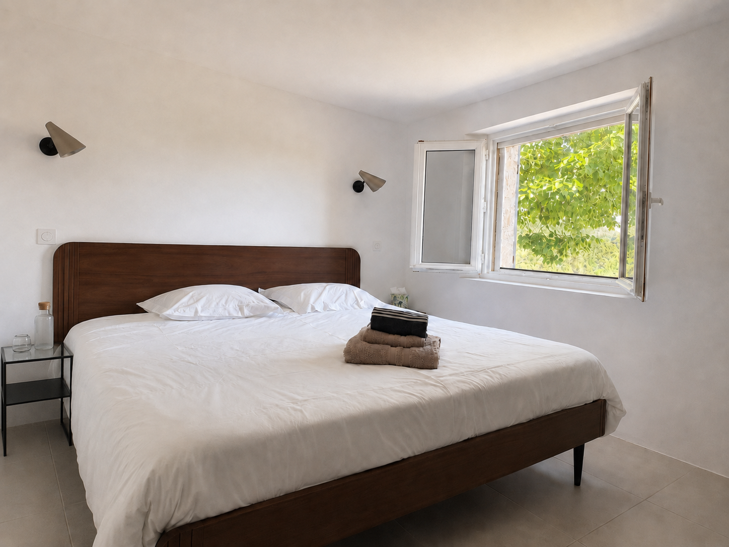 A minimalist bedroom with a large bed, white bedding, and towels on top. Two black sconces on the white wall, an open window with green foliage outside, and a small black bedside table with a glass and a bottle.
