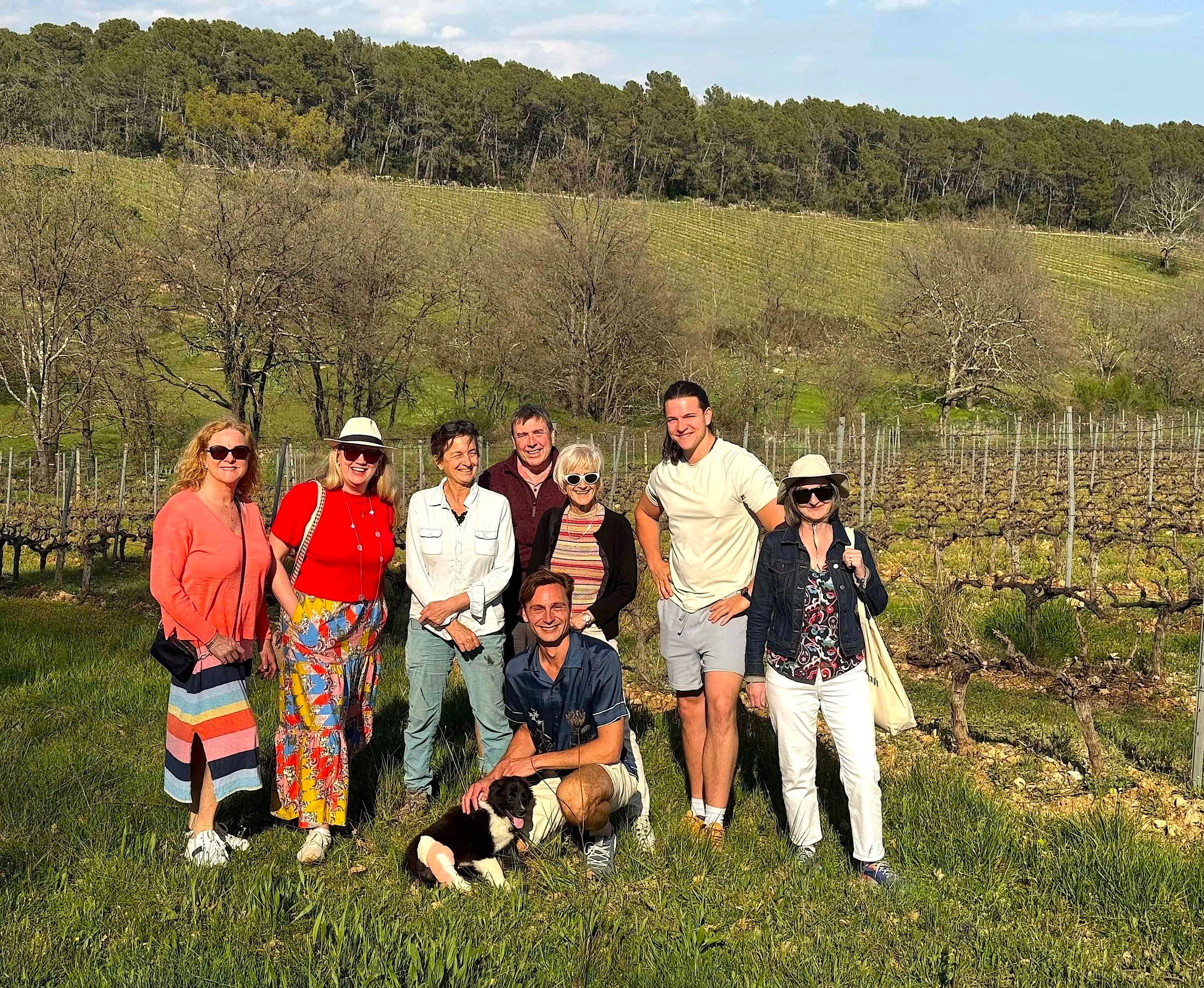 A group of ten people, including men and women, standing outdoors in a vineyard with rolling hills and trees in the background. They are smiling and dressed casually, with one person holding a black and white dog.