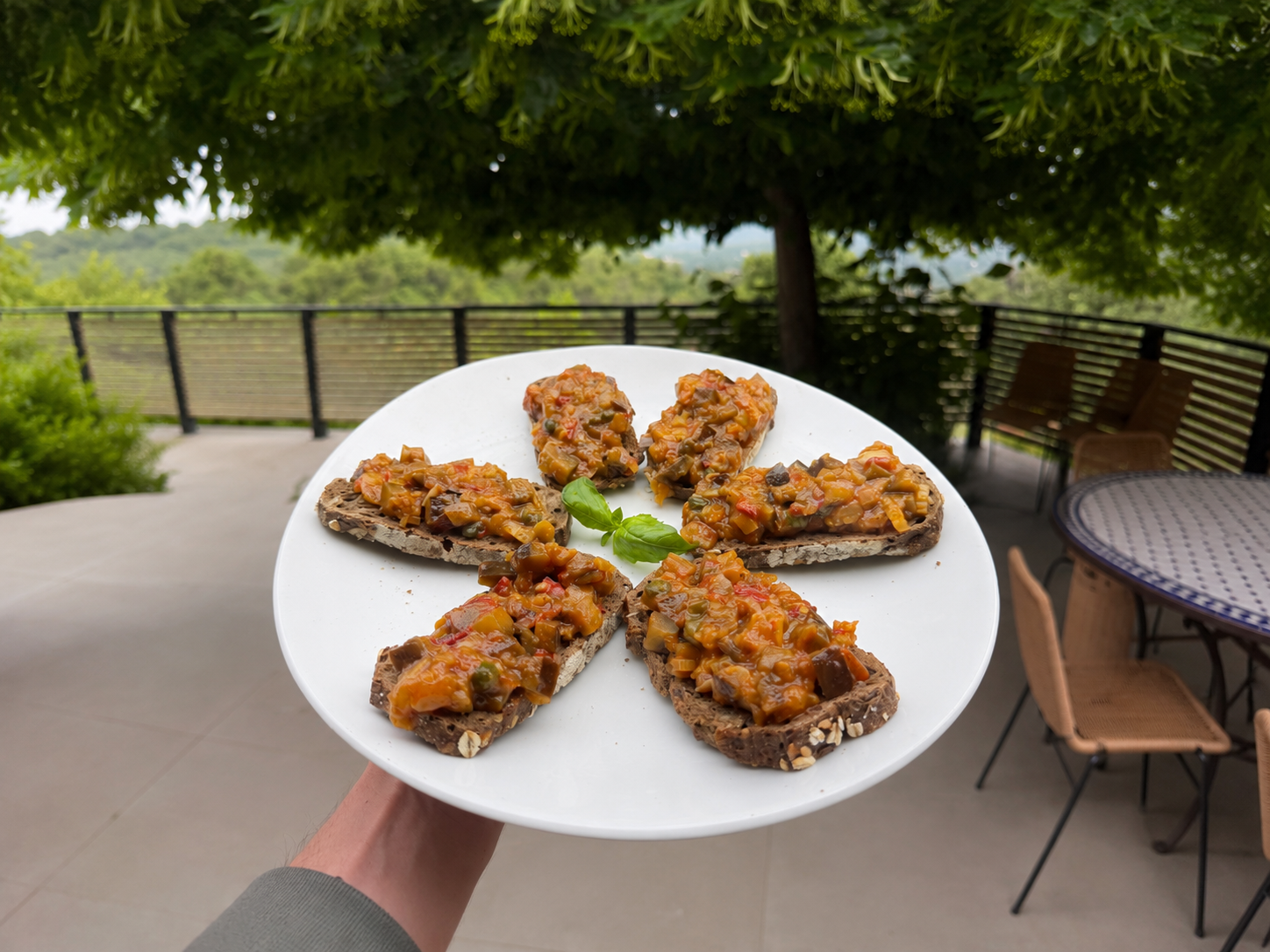 Plate with five slices of bread topped with vegetable chutney, garnished with a small basil leaf, held outdoors with a patio and trees in the background.