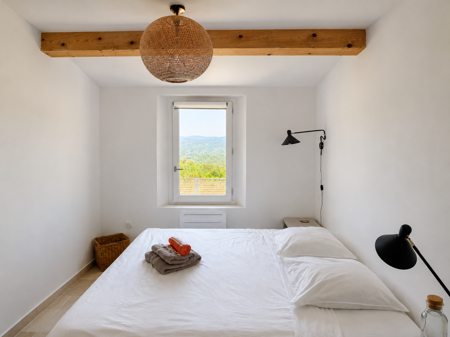 A minimalist bedroom with white walls, a window showing a scenic landscape, a bed with white bedding, a black wall-mounted lamp, a small side table, a floor lamp, a wicker basket, and neatly folded towels on the bed. A wooden beam hangs from the ceiling.