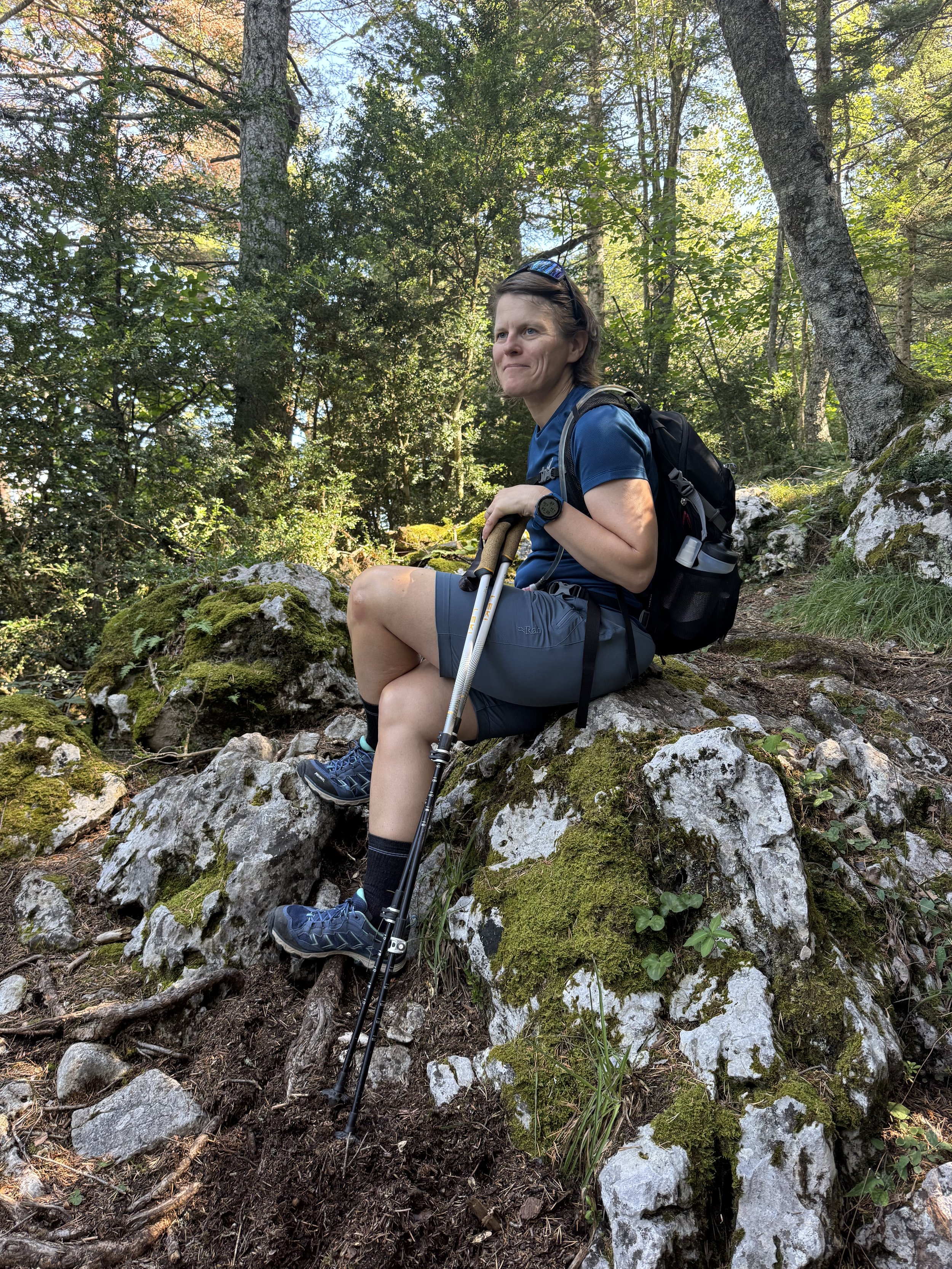 A woman sitting on moss-covered rocks in a forest during a hiking trip, holding hiking poles, wearing a blue shirt, gray shorts, hiking shoes, and a backpack.
