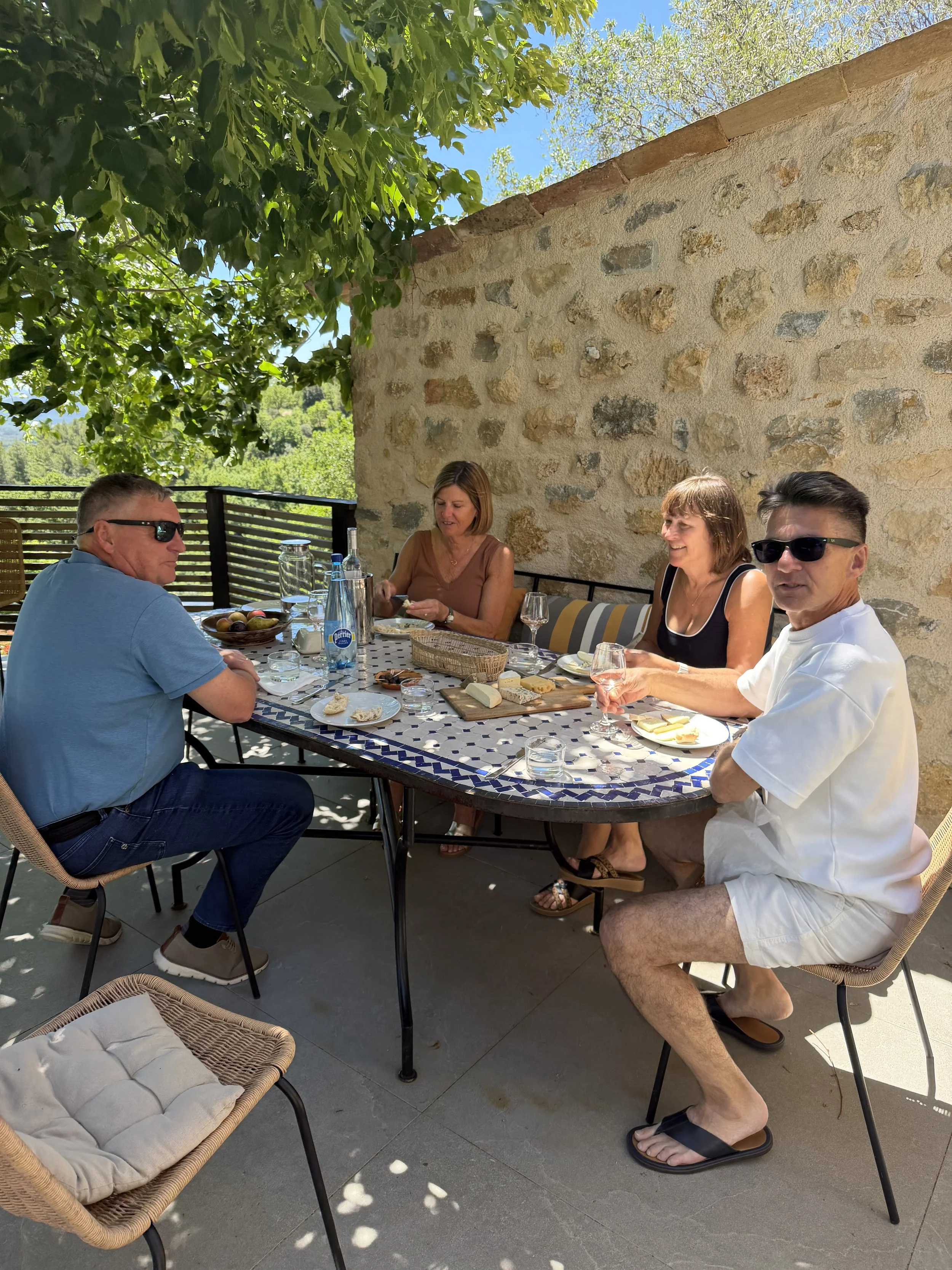 Four people sitting around a dining table outdoors with a stone wall and green trees in the background. They are enjoying a meal with cheese, bread, wine, and bottled water.