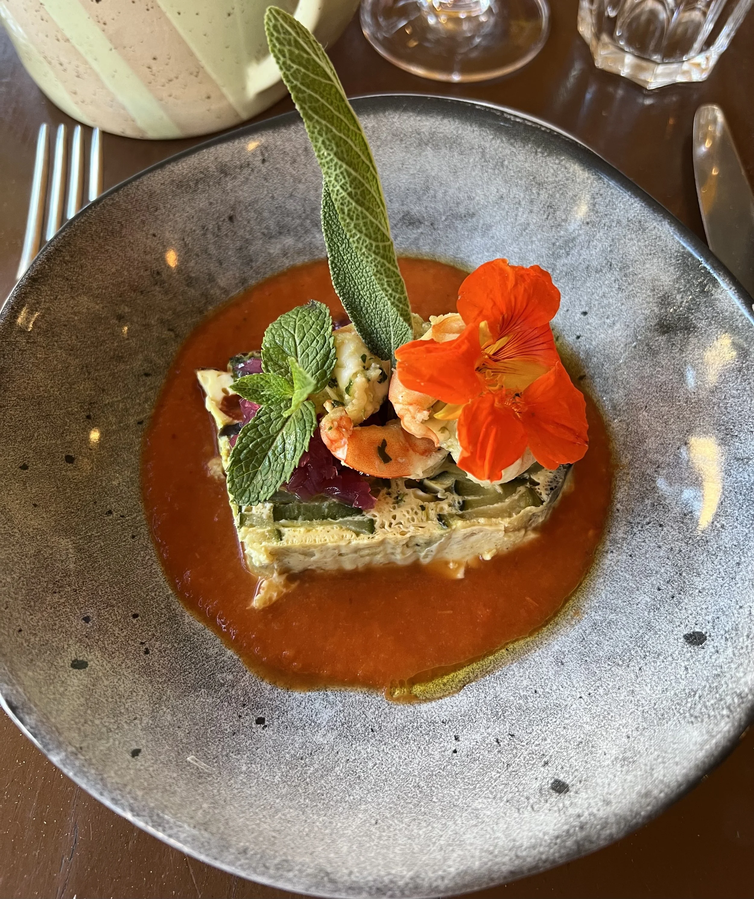 A plated gourmet dish with a vegetable terrine topped with herbs and edible flowers, served in a red sauce on a dark textured plate.