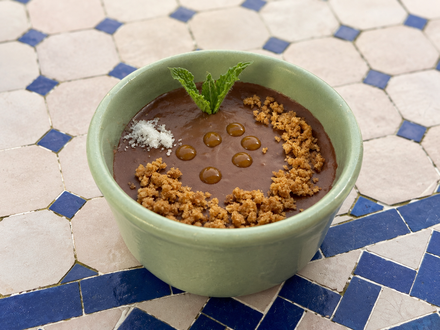 Chocolate mousse garnished with coconut, crushed cookies, mint leaves, and caramel dots on a green bowl placed on a tiled table.