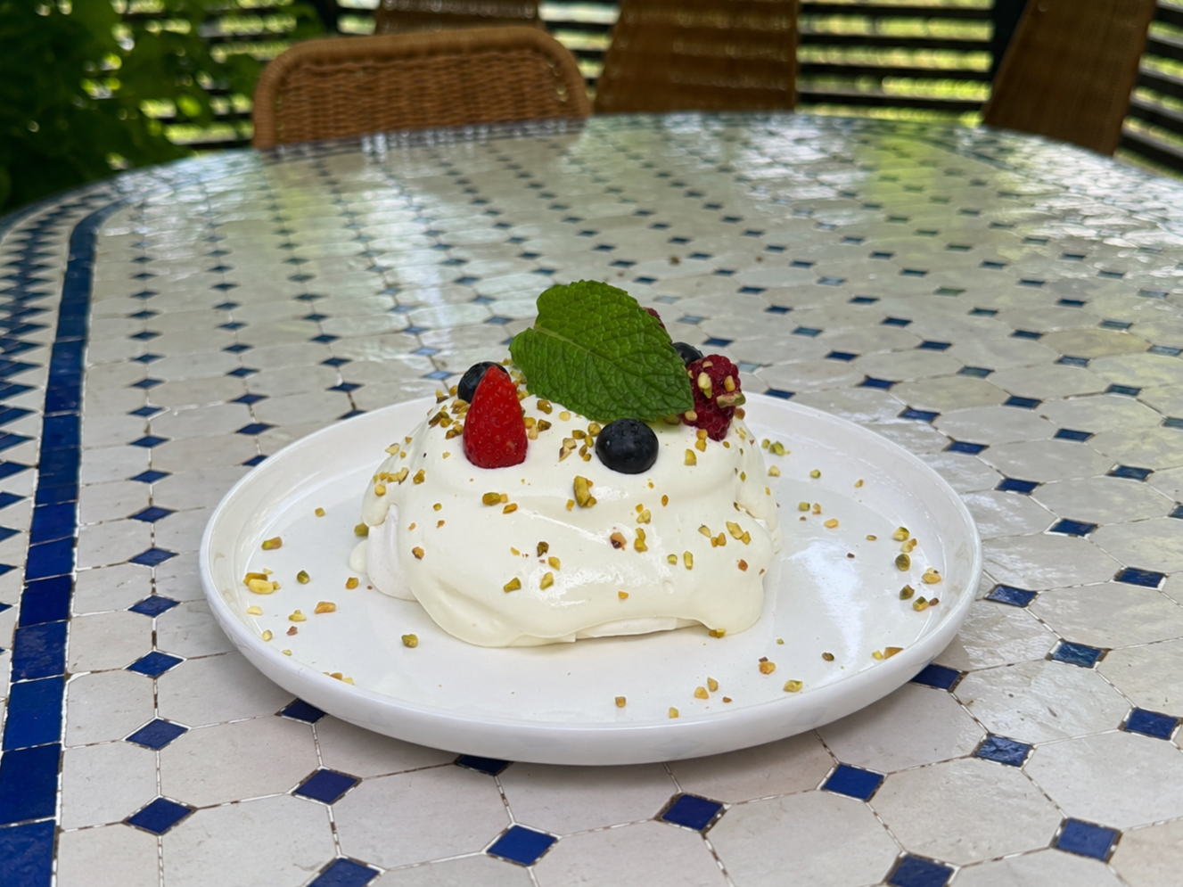 Meringue dessert topped with fresh berries, mint leaves, and chopped pistachios on a white plate outside.