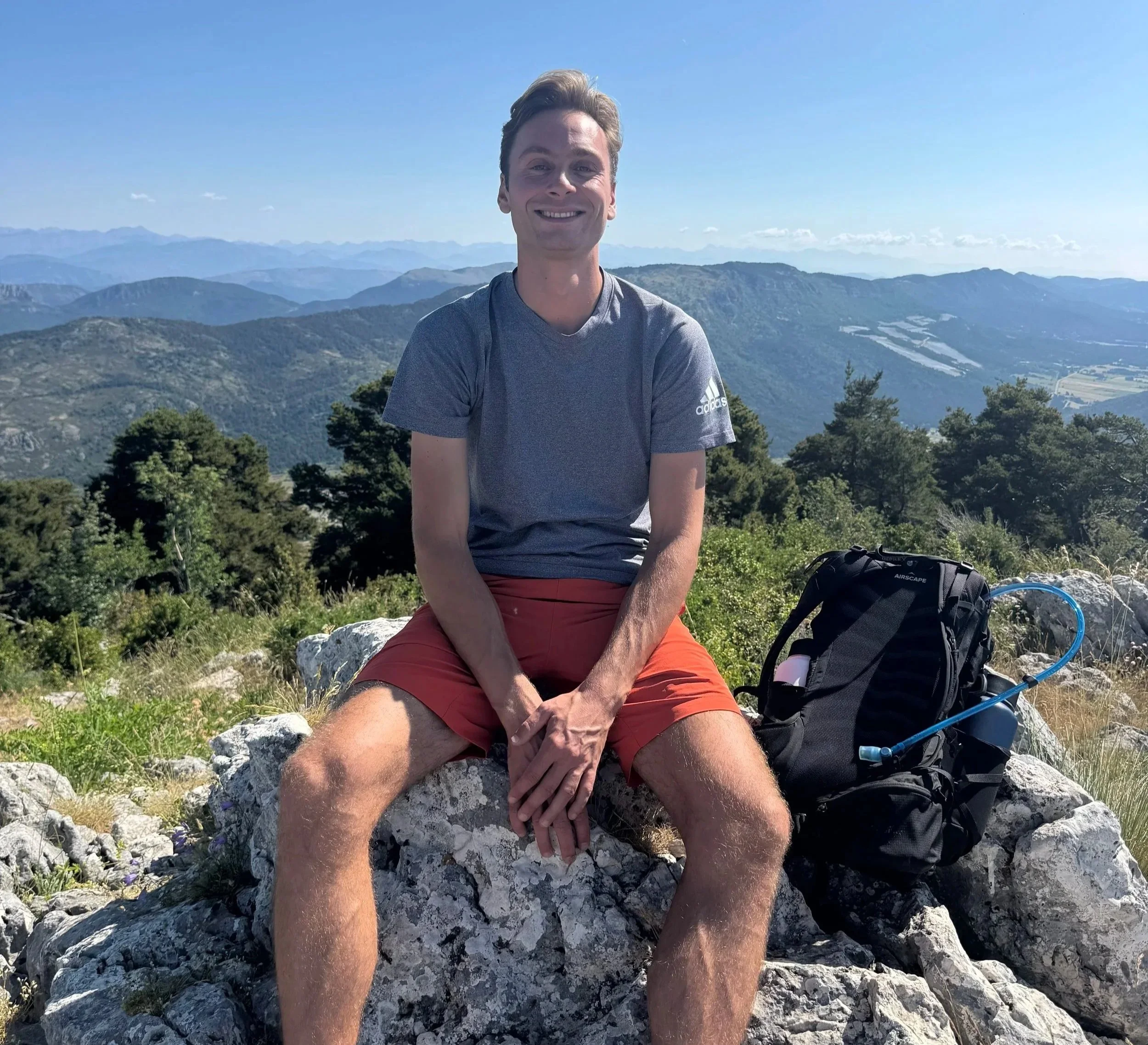 A young man sitting on rocks in a mountainous landscape, smiling at the camera, with a backpack on the ground next to him, under a clear blue sky with distant mountain ranges.