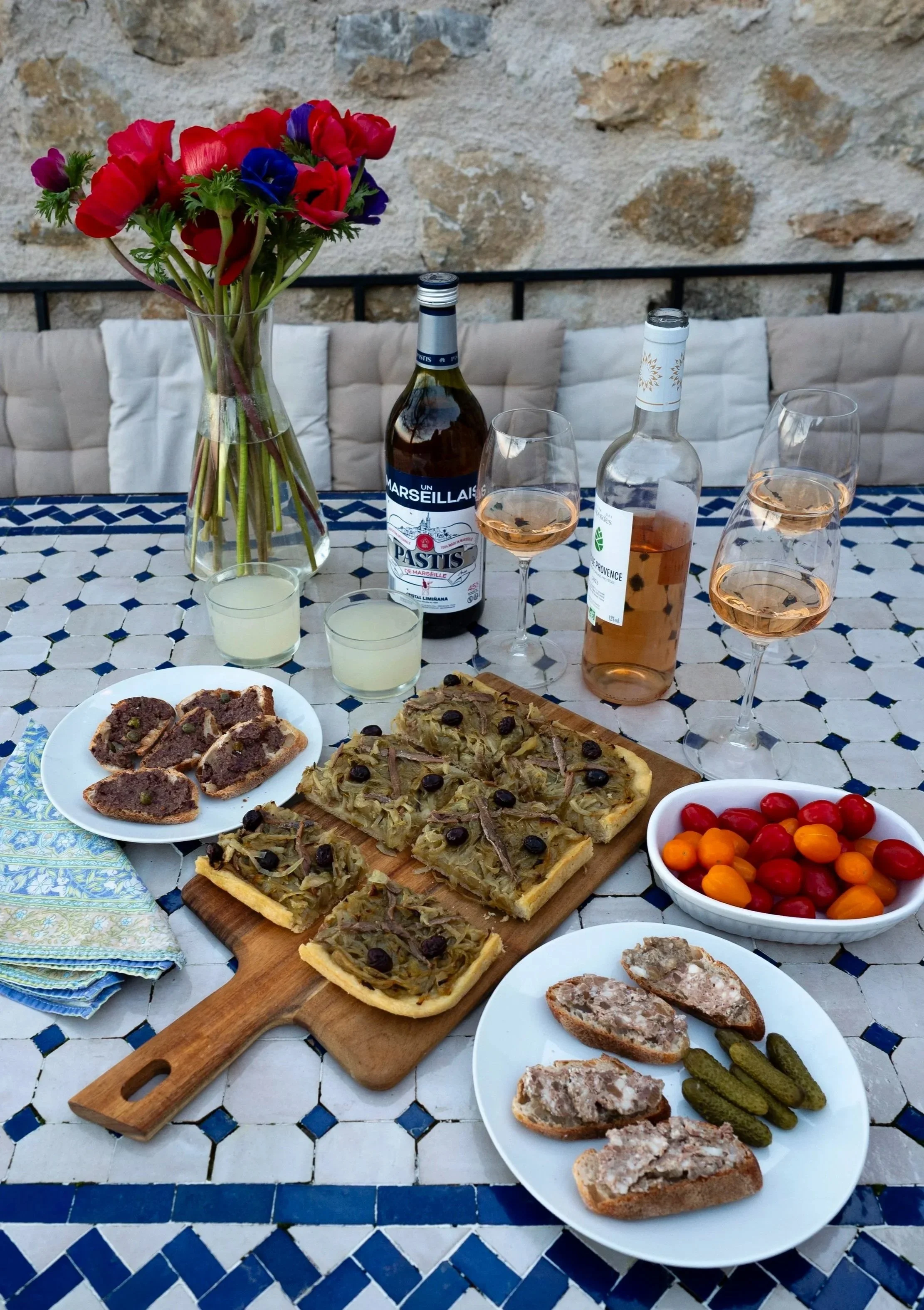 Table with various foods including sliced bread with meat spread, vegetable flan, cherry tomatoes, and snacks; a vase with colorful flowers; bottles of rosé wine and sparkling water; glasses of rosé wine and water; a plate of cherries; napkins, and a stone wall in the background.