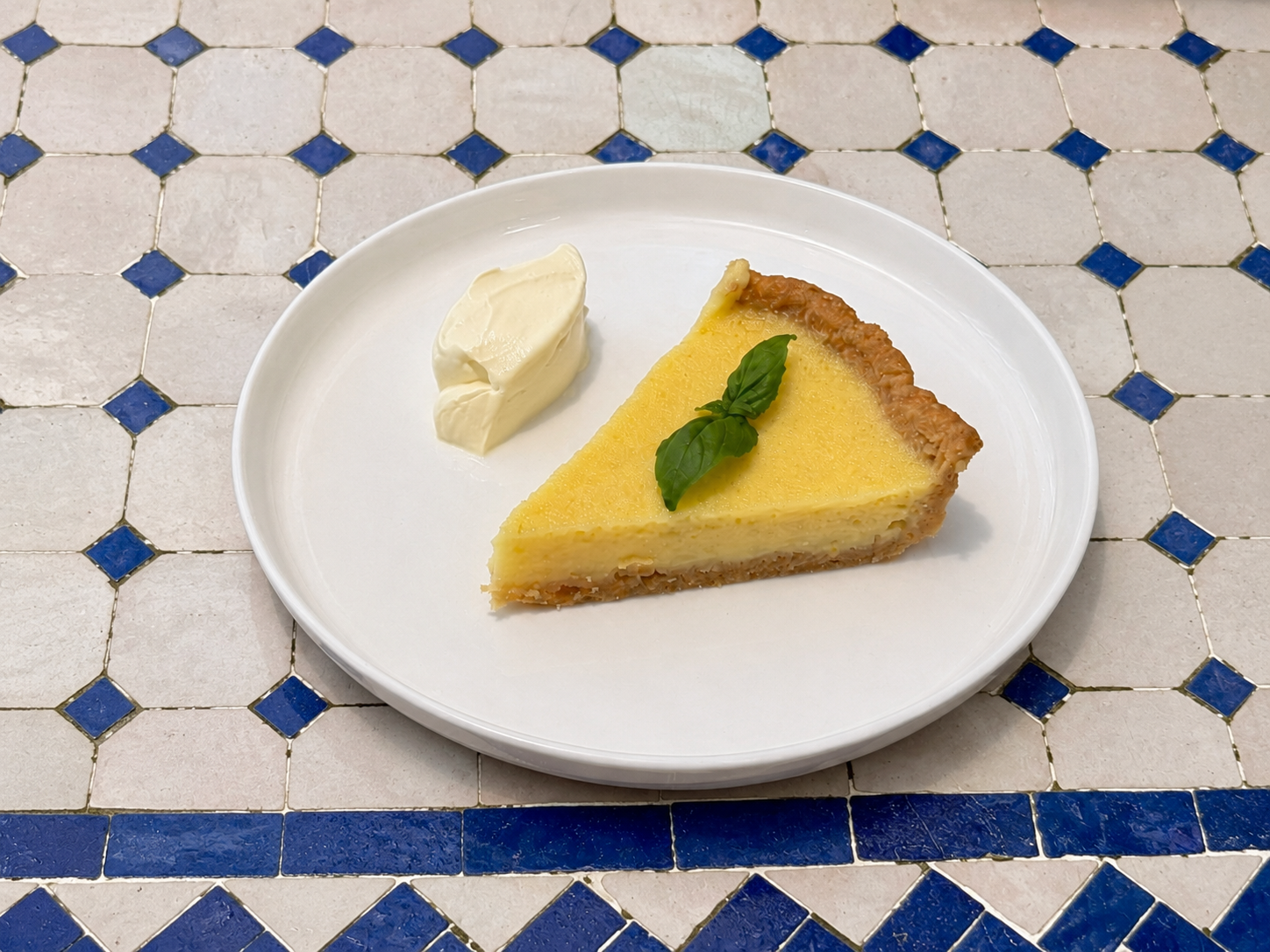 Slice of lemon tart with basil leaves and a dollop of whipped cream on a white plate, on a tiled table.