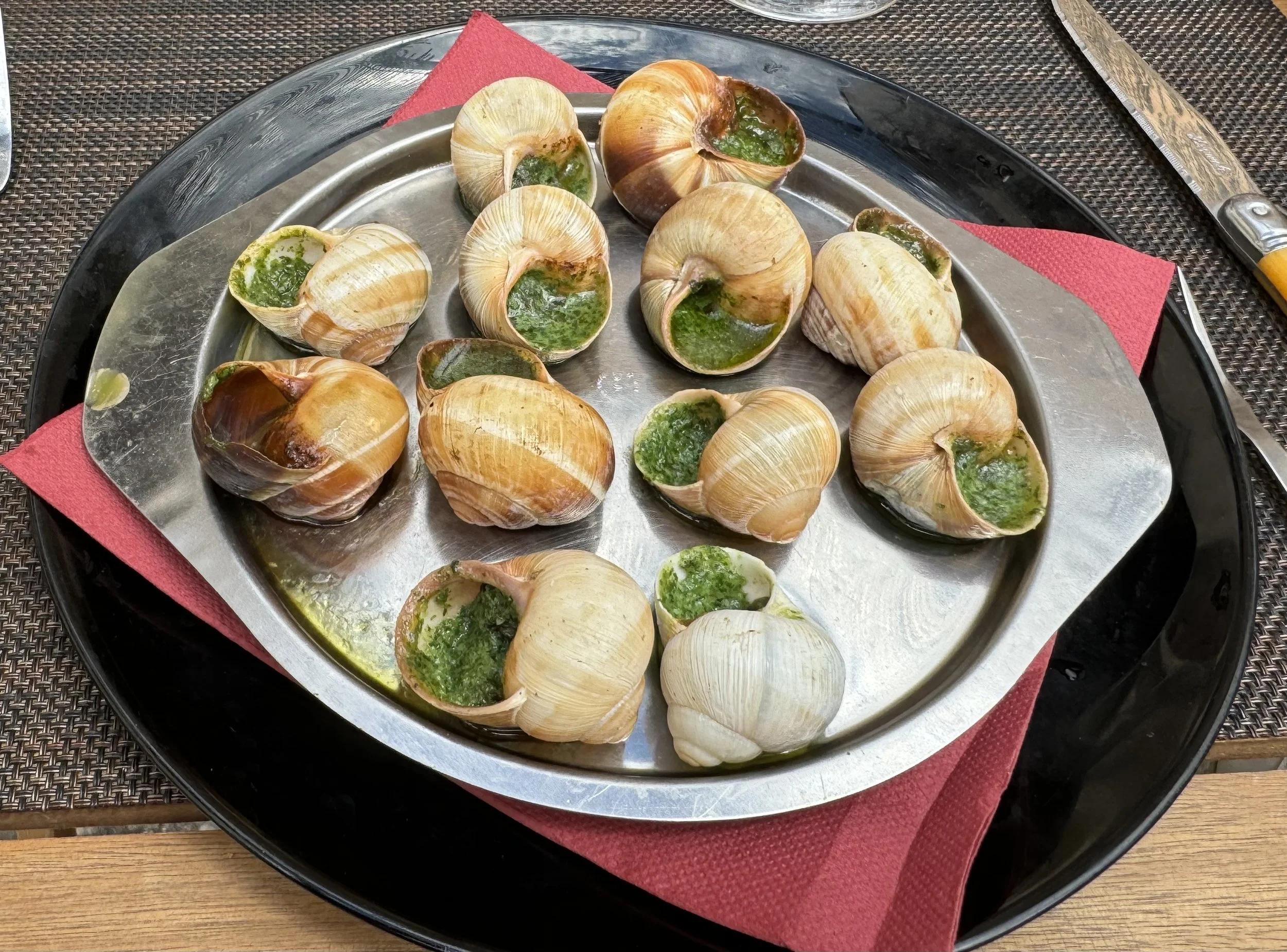Close-up of stuffed escargot shells with green herb butter on a silver platter, on a table with red napkin and cutlery.