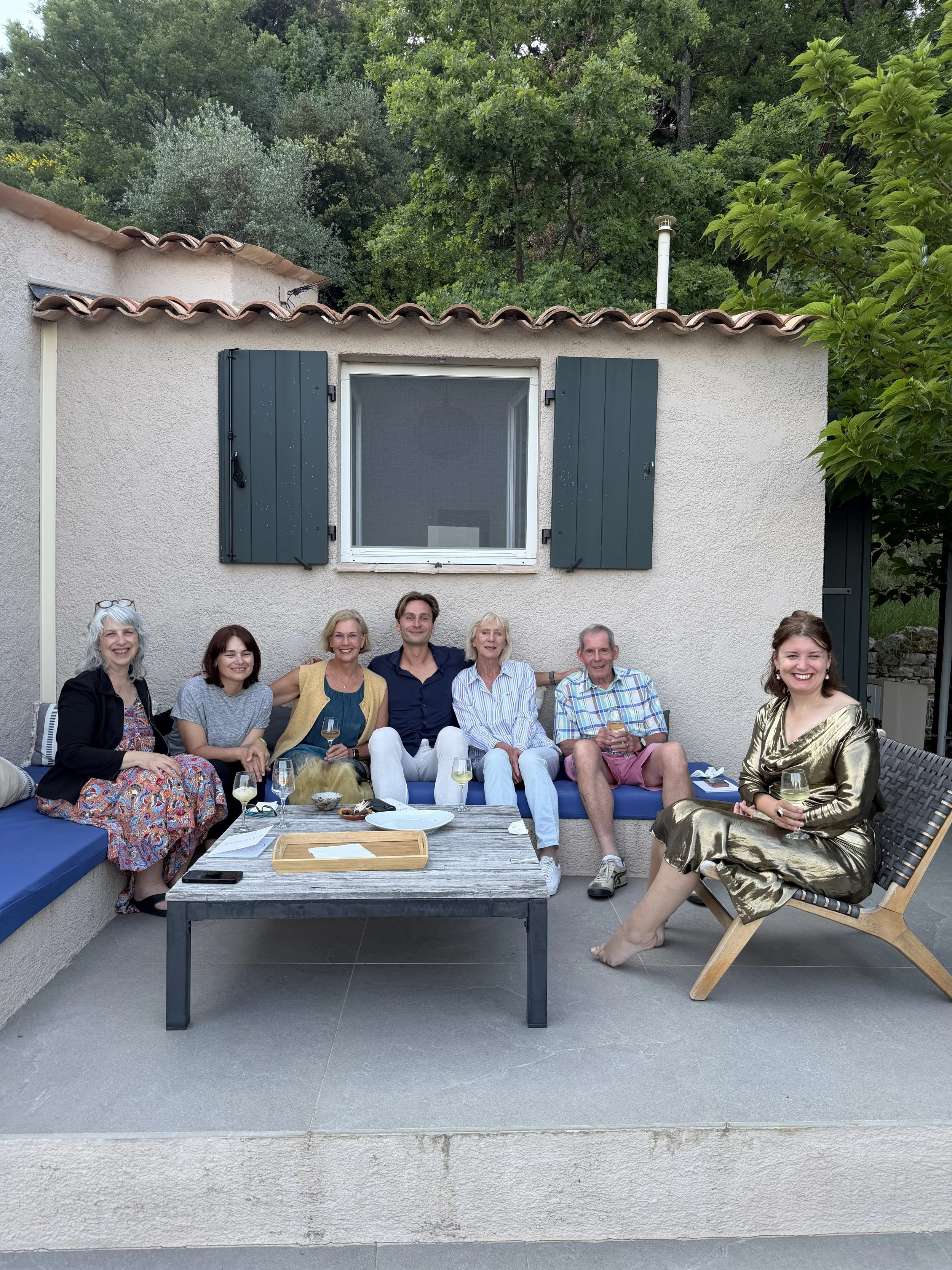 A group of people sitting and smiling on a patio in front of a beige wall with a window and dark green shutters, surrounded by lush greenery and trees.