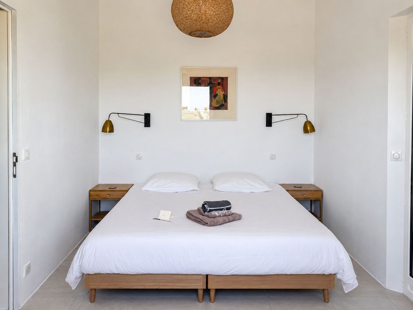 A neatly made bed with white linens and two pillows, flanked by two wooden bedside tables with small black remote controls. Above the bed are two black and brass wall-mounted reading lamps, and a framed abstract artwork hanging on a white wall. On the bed are folded towels and a note.