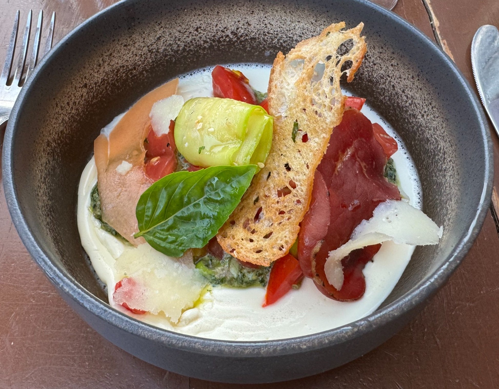 Bowl of fresh Caprese salad with tomatoes, mozzarella, basil, parmesan, pesto, and a crispy bread chip.