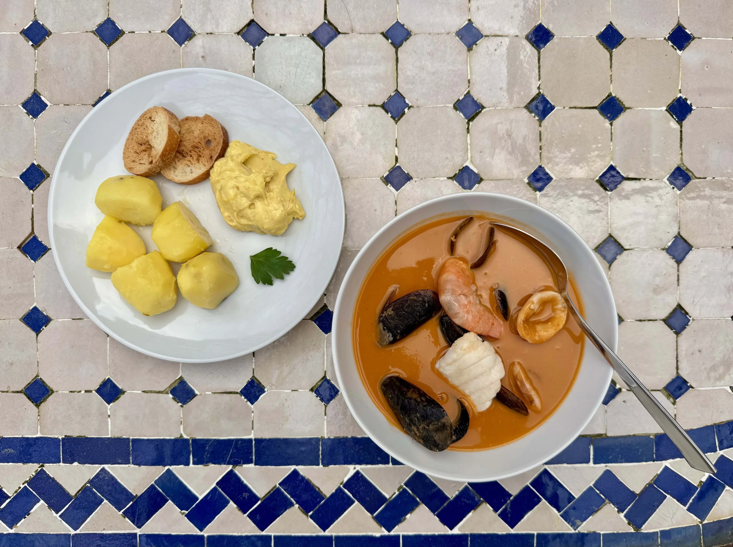 A white plate of peeled boiled potatoes, slices of bread, and a dollop of rouille; next to it a bowl of seafood soup, bouillabaisse with prawns, mussels, and squid, placed on a tiled surface with a mix of white, and blue tiles.