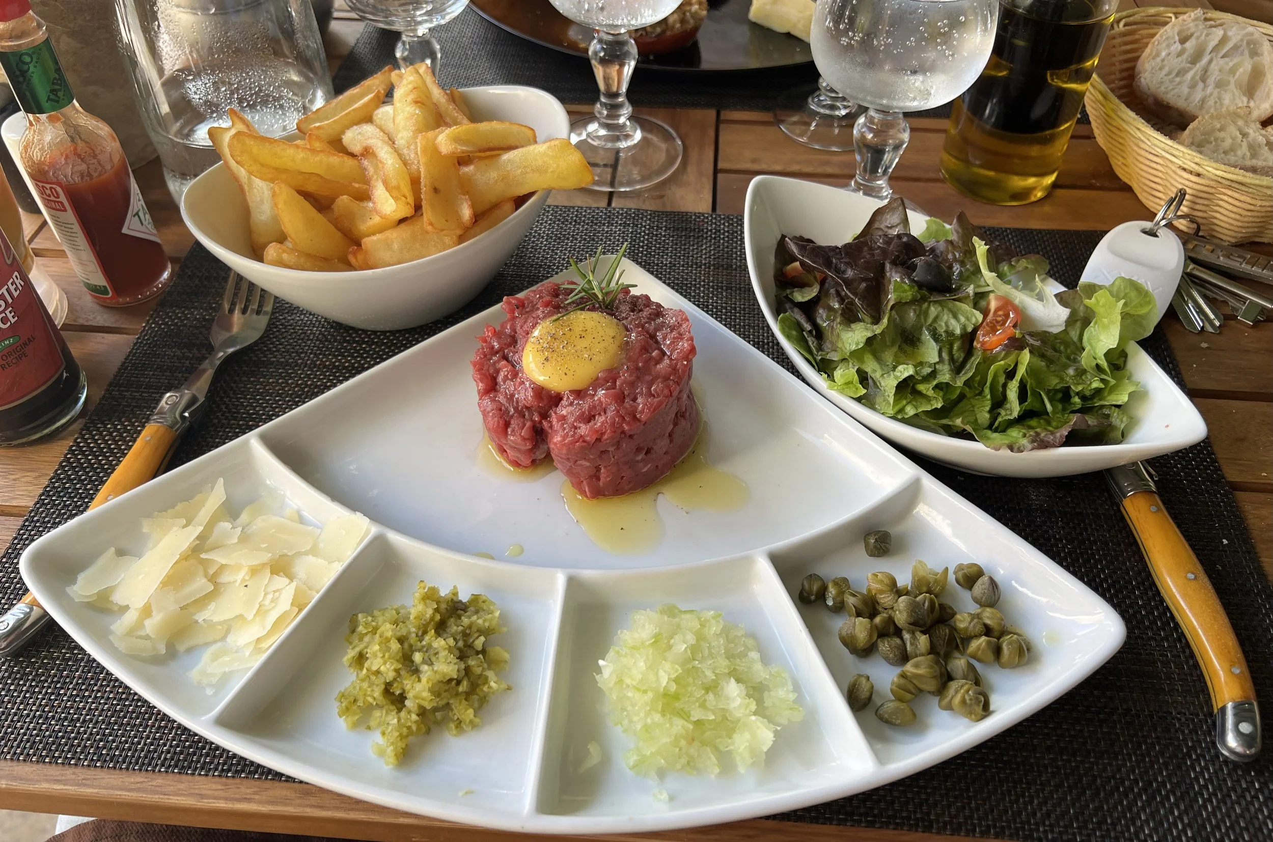 Steak tartare with a raw egg yolk on top, served with grated cheese, chopped pickles, and capers on a white plate, with a side of salad and French fries on a table with cutlery and condiments.