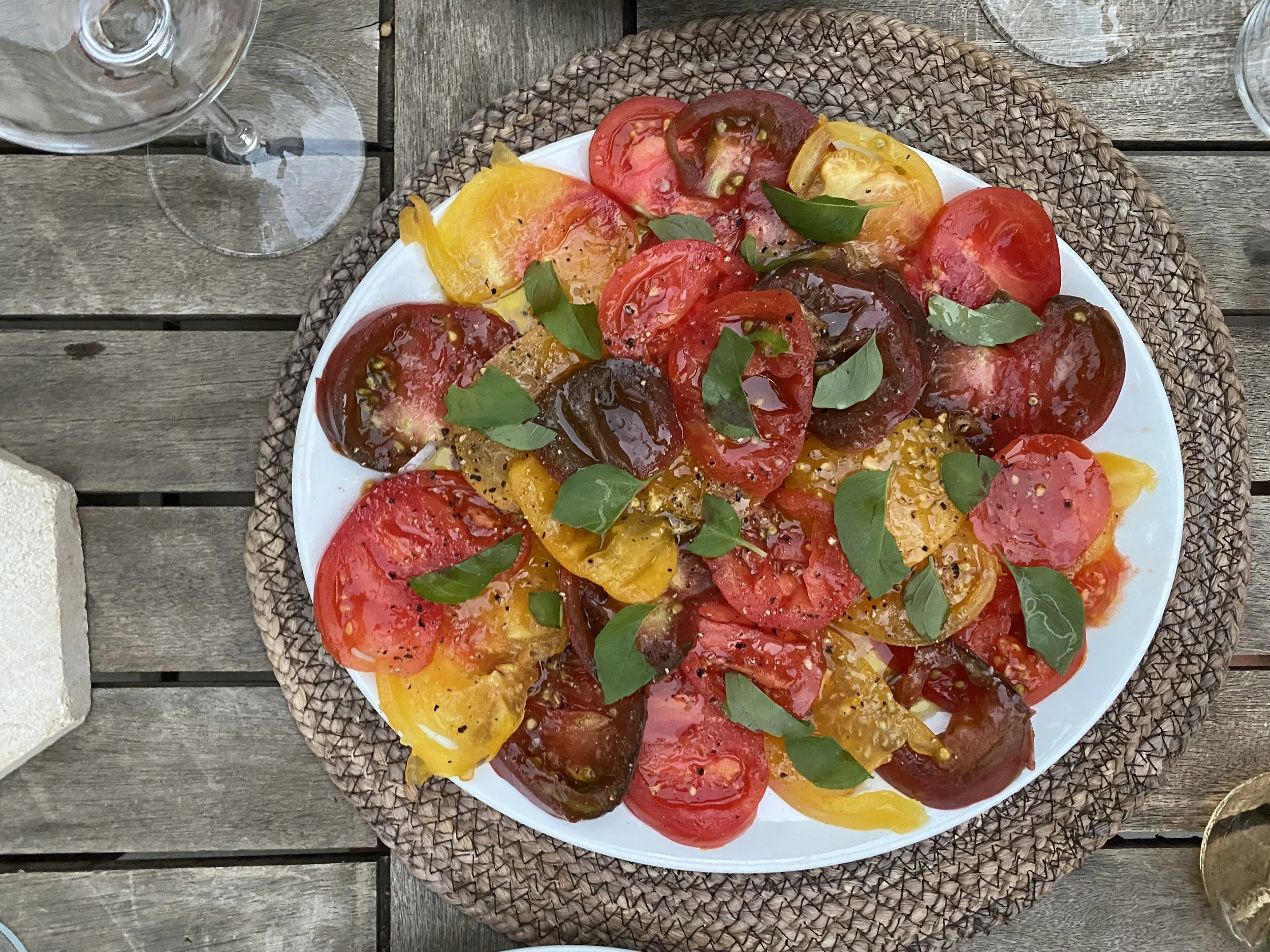 A plate of sliced heirloom tomatoes with basil leaves on top, served on a woven placemat on a wooden table, with wine glasses nearby.