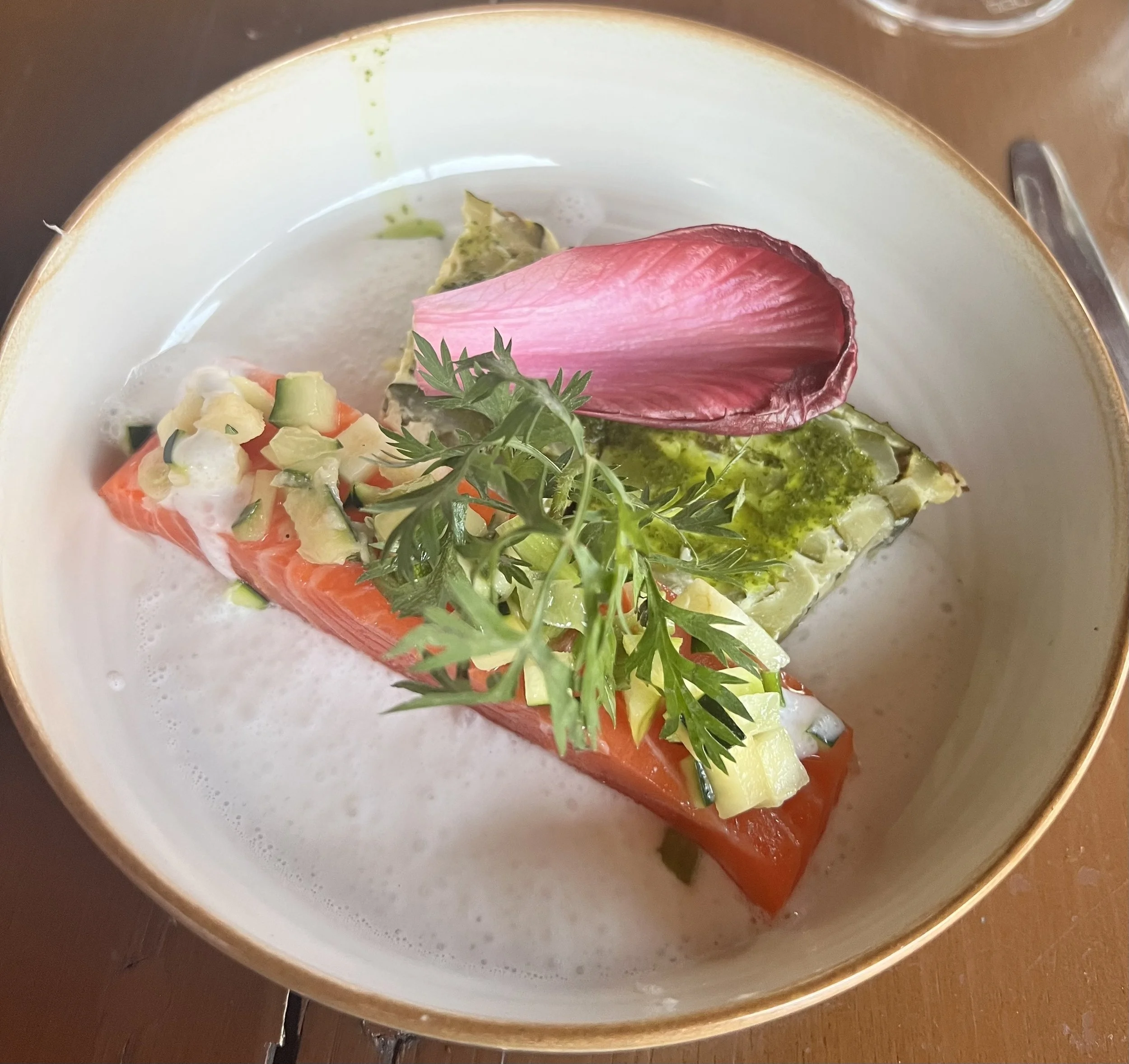 A gourmet dish featuring cooked salmon topped with diced vegetables, garnished with fresh herbs, a single pink petal, and a green sauce on a white plate with a gold rim.