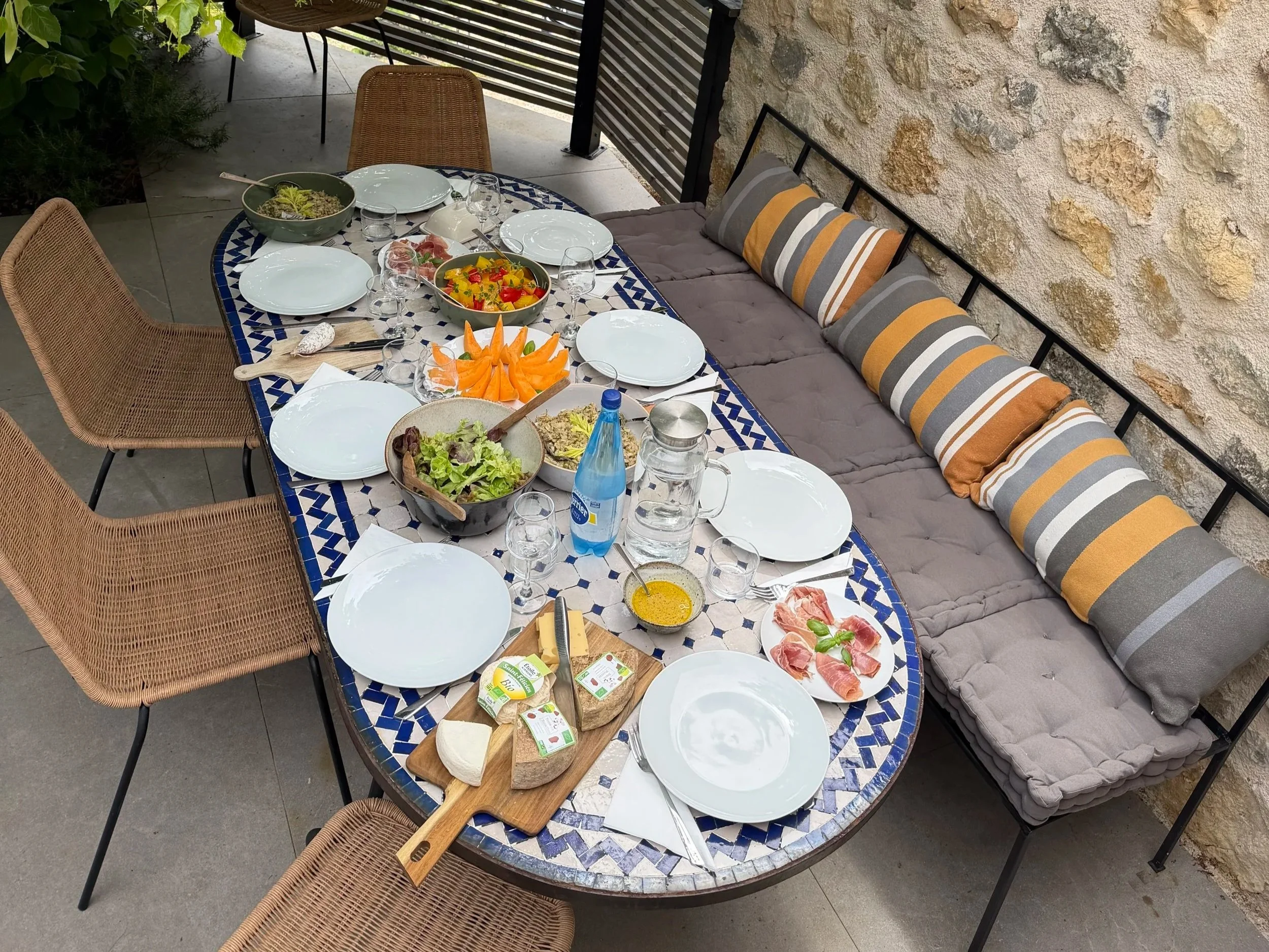 A table set for a meal with plates, glasses, and bowls of food, including salad and sliced cantaloupe, in an outdoor patio with wicker chairs and decorative pillows on a bench against a textured wall.