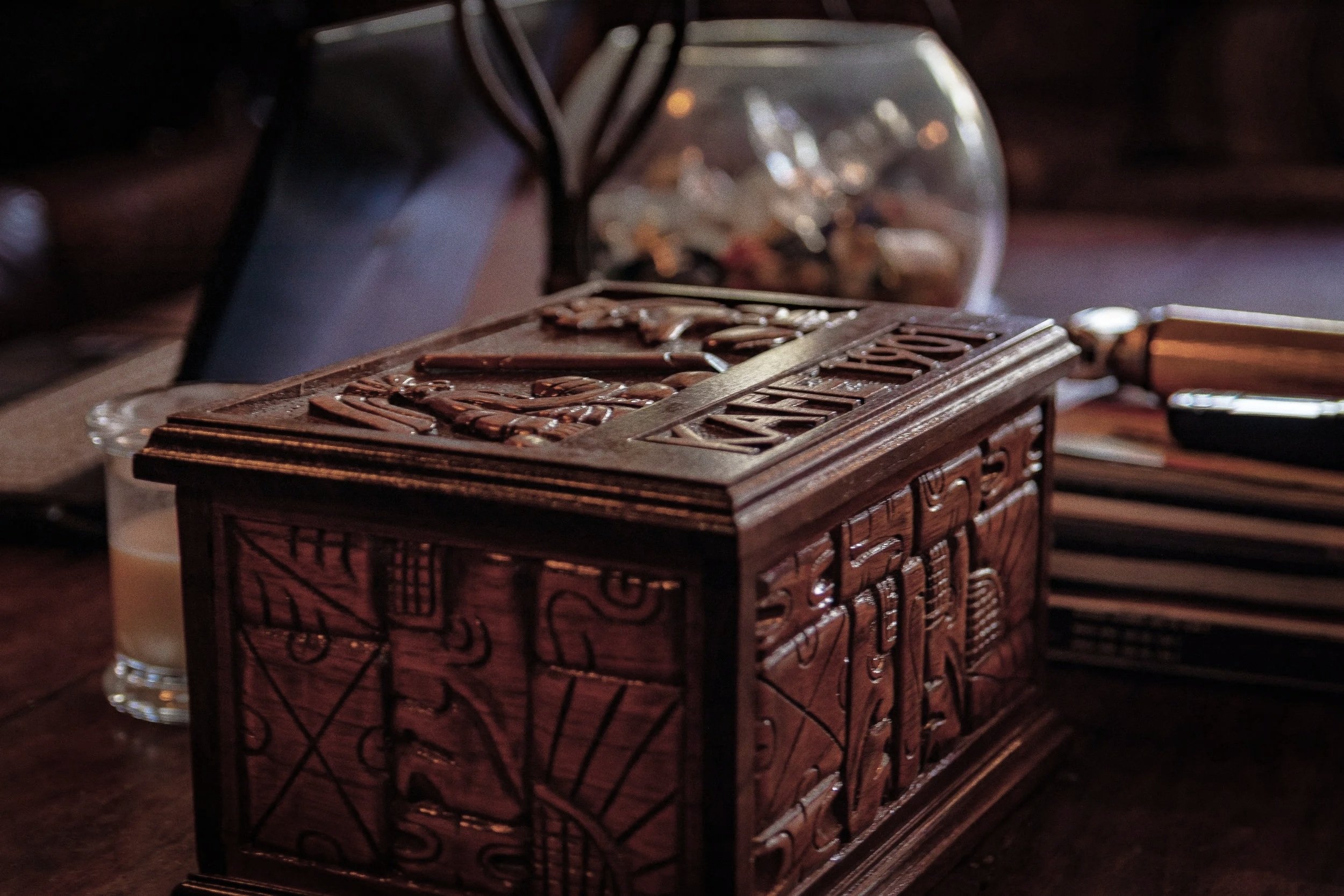 A wooden decorative box with carved designs on a table, with a glass of coffee, a partially visible pen, and a glass container with pills or small objects in the background.