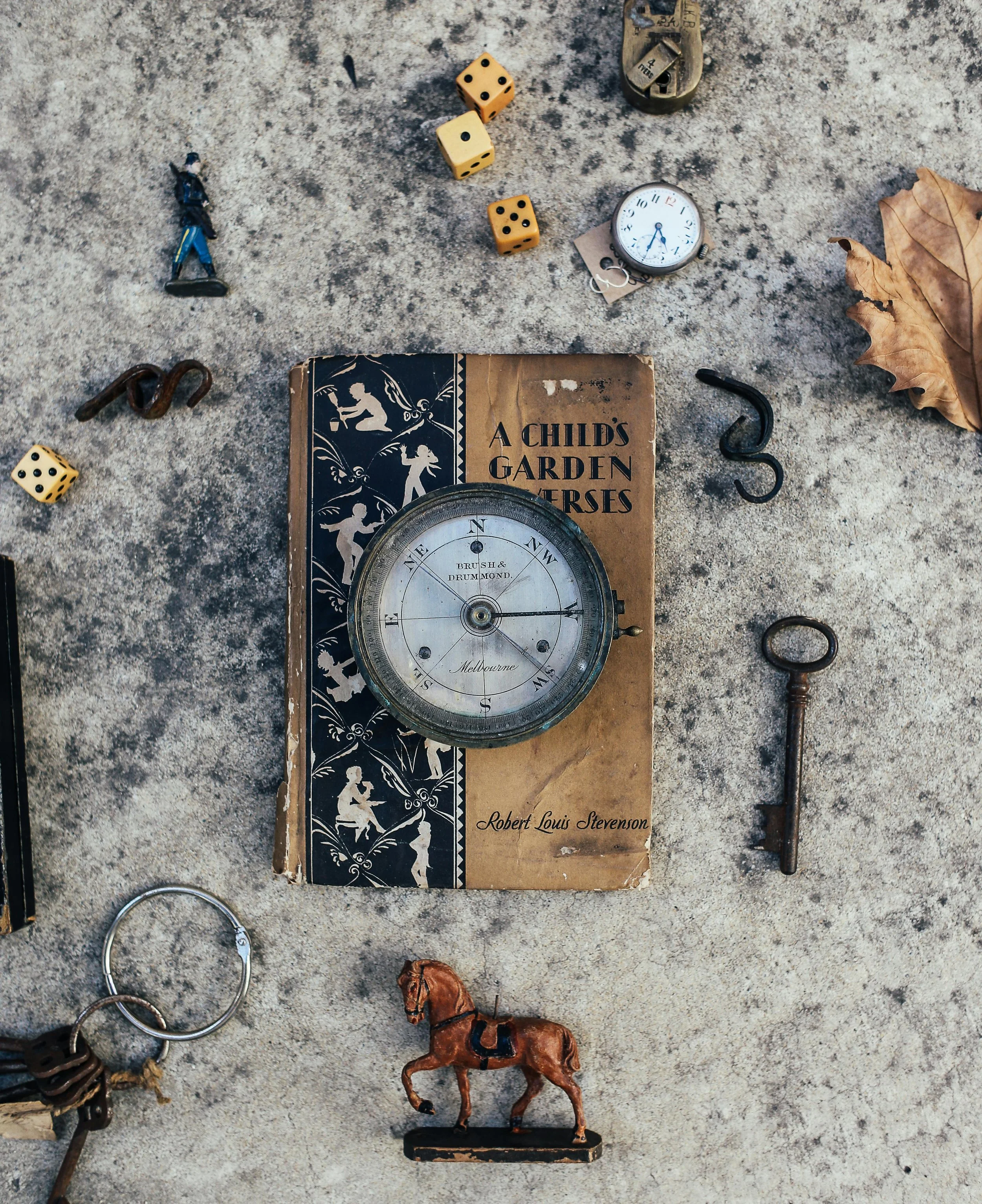 A collection of vintage objects on a concrete surface, including a book titled 'A Child's Garden of Verses,' a compass, a key, a horse figurine, a pocket watch, a few dice, a small soldier figurine, a dried leaf, and various small metal objects.