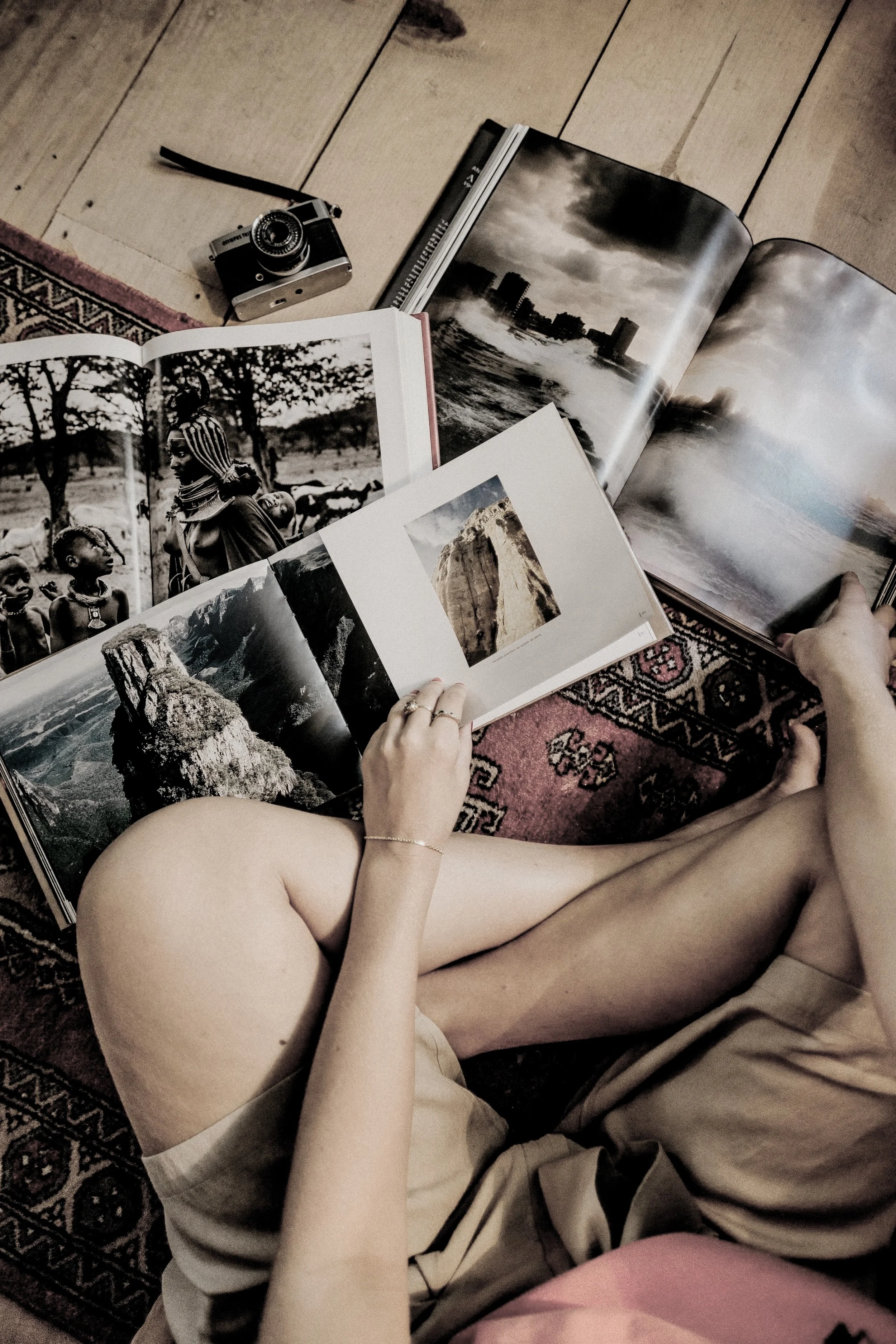 A person sitting on a patterned carpet looking through open travel books, with a small camera and closed book nearby.