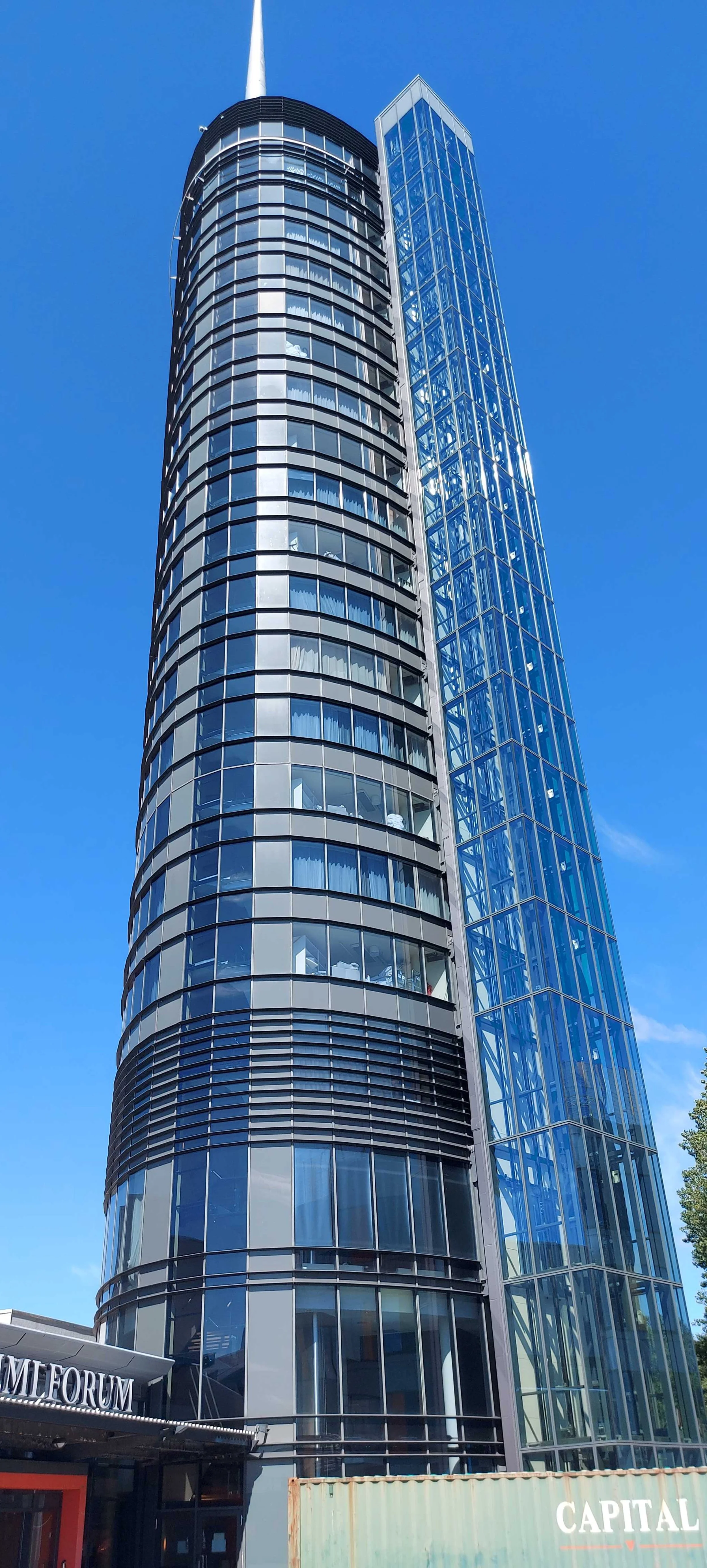 To høye, moderne skyskrapere med glassfasade mot en klar blå himmel, en har en rund form og den andre er kvadratisk.